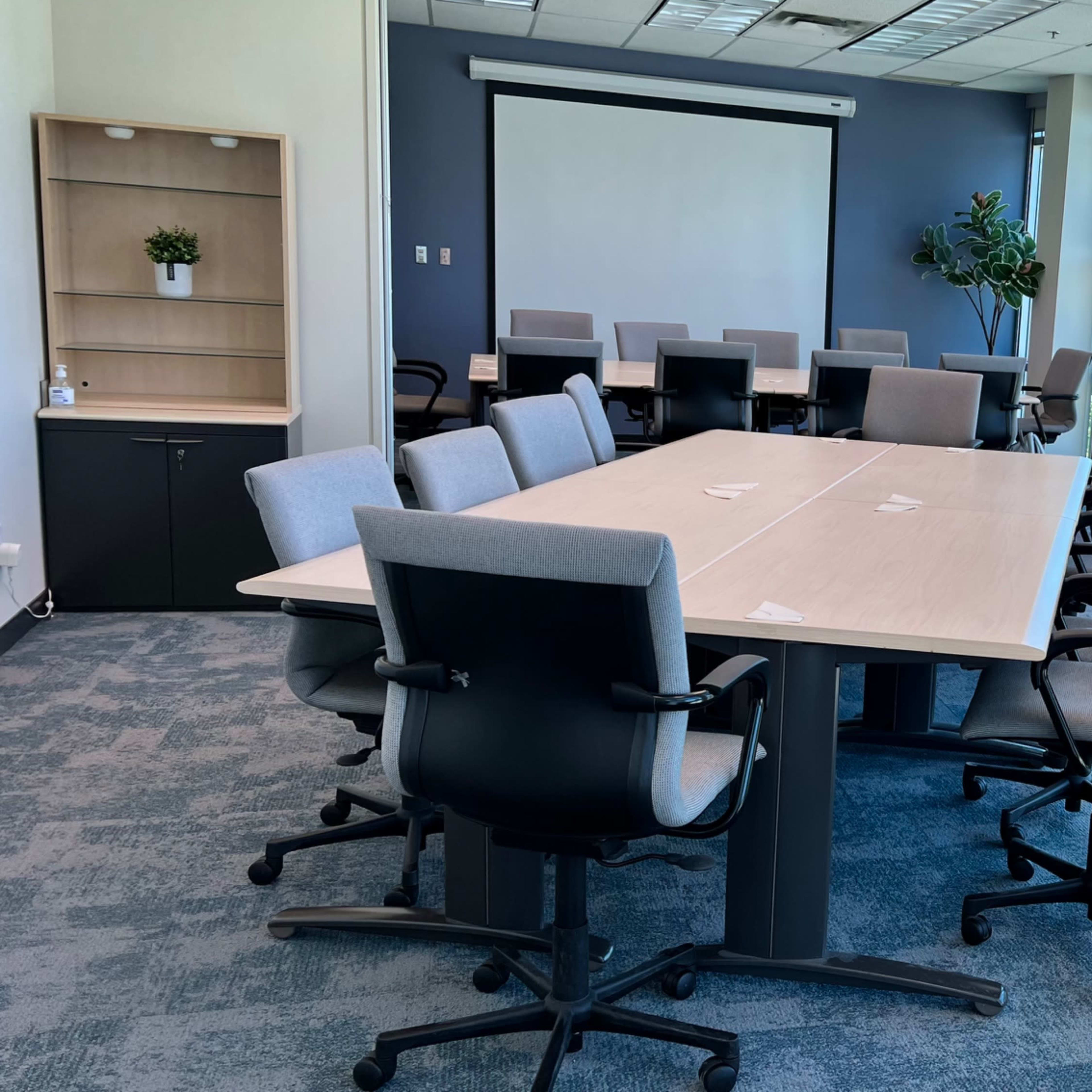 A spacious conference room features a large table surrounded by ergonomic chairs, with a projector screen and windows that provide ample natural light.