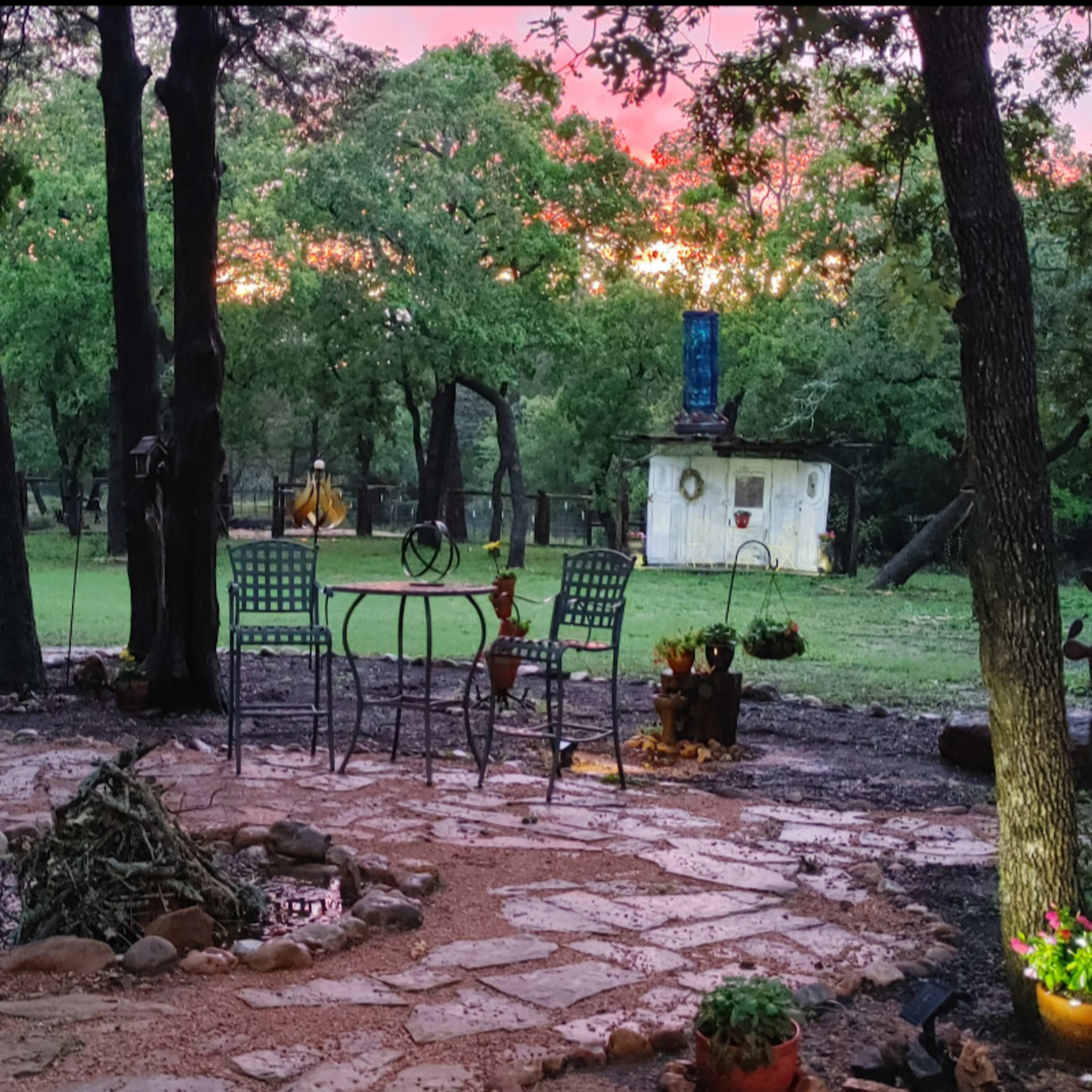 A circular stone fire pit is surrounded by a patio area with a table and chairs, set against a backdrop of trees and a rustic shed under a colorful sunset sky.