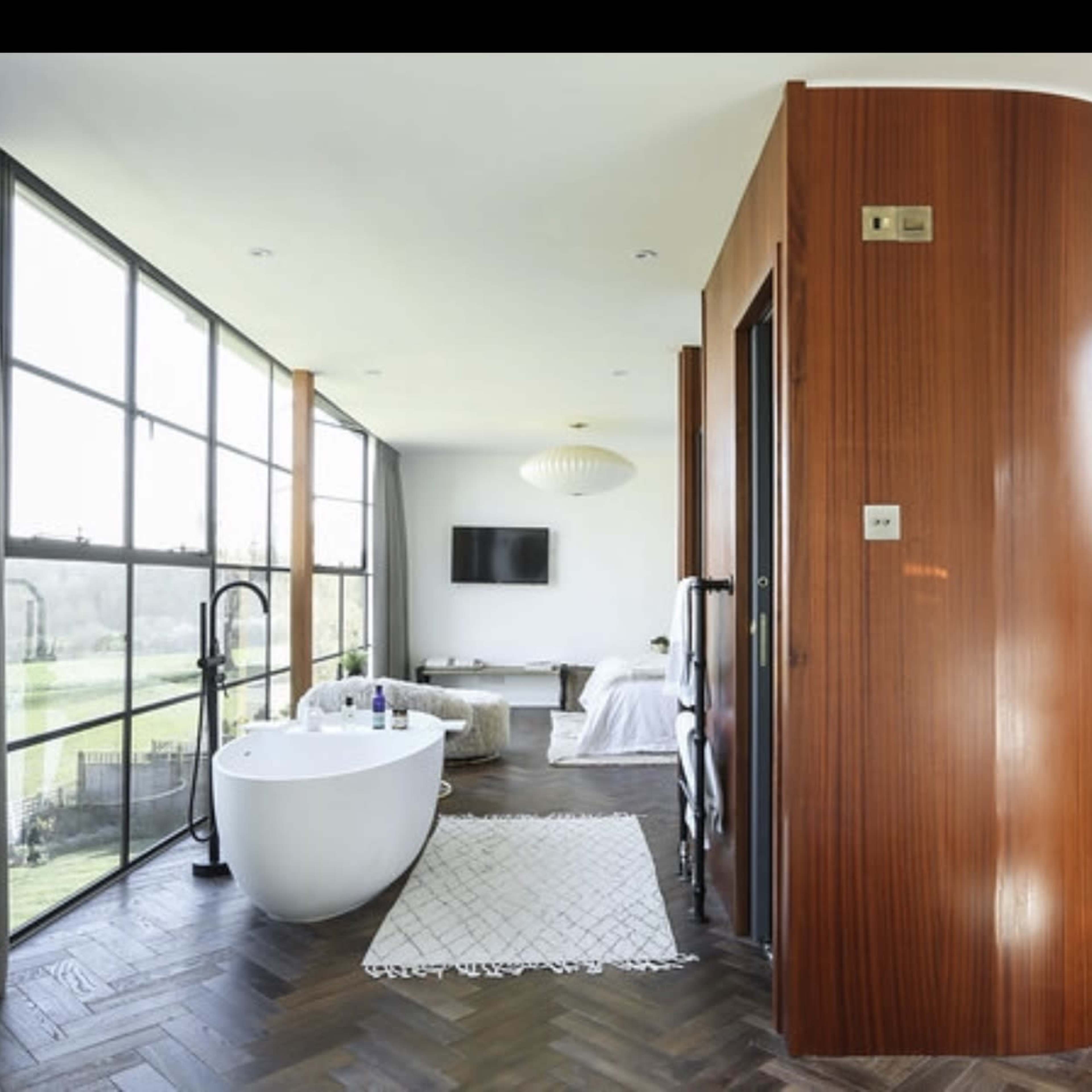 A modern, open-concept bathroom and bedroom area with large windows, a freestanding bathtub, and wooden accents.