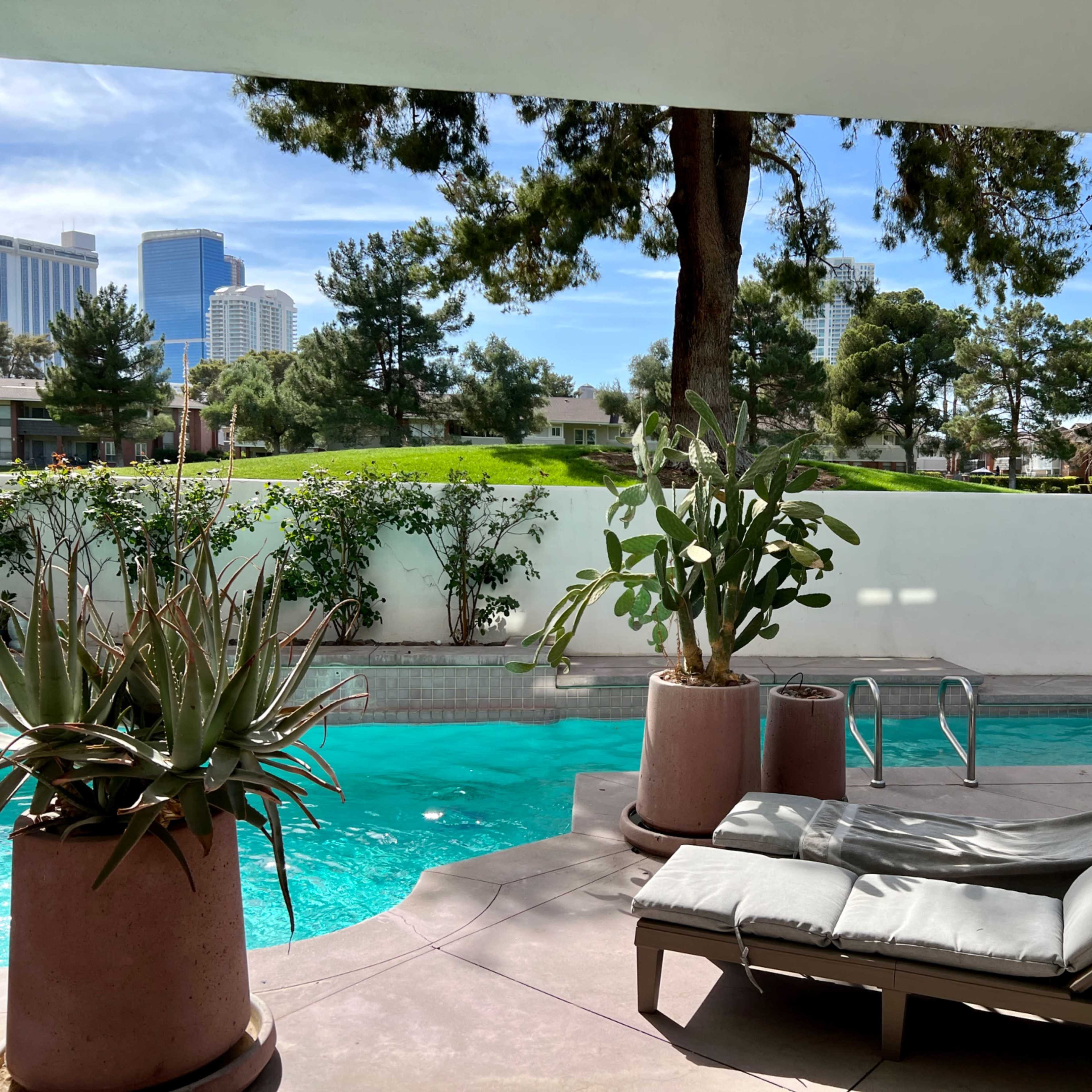 A swimming pool with a lounge chair beside it, surrounded by greenery and overlooking a city skyline.
