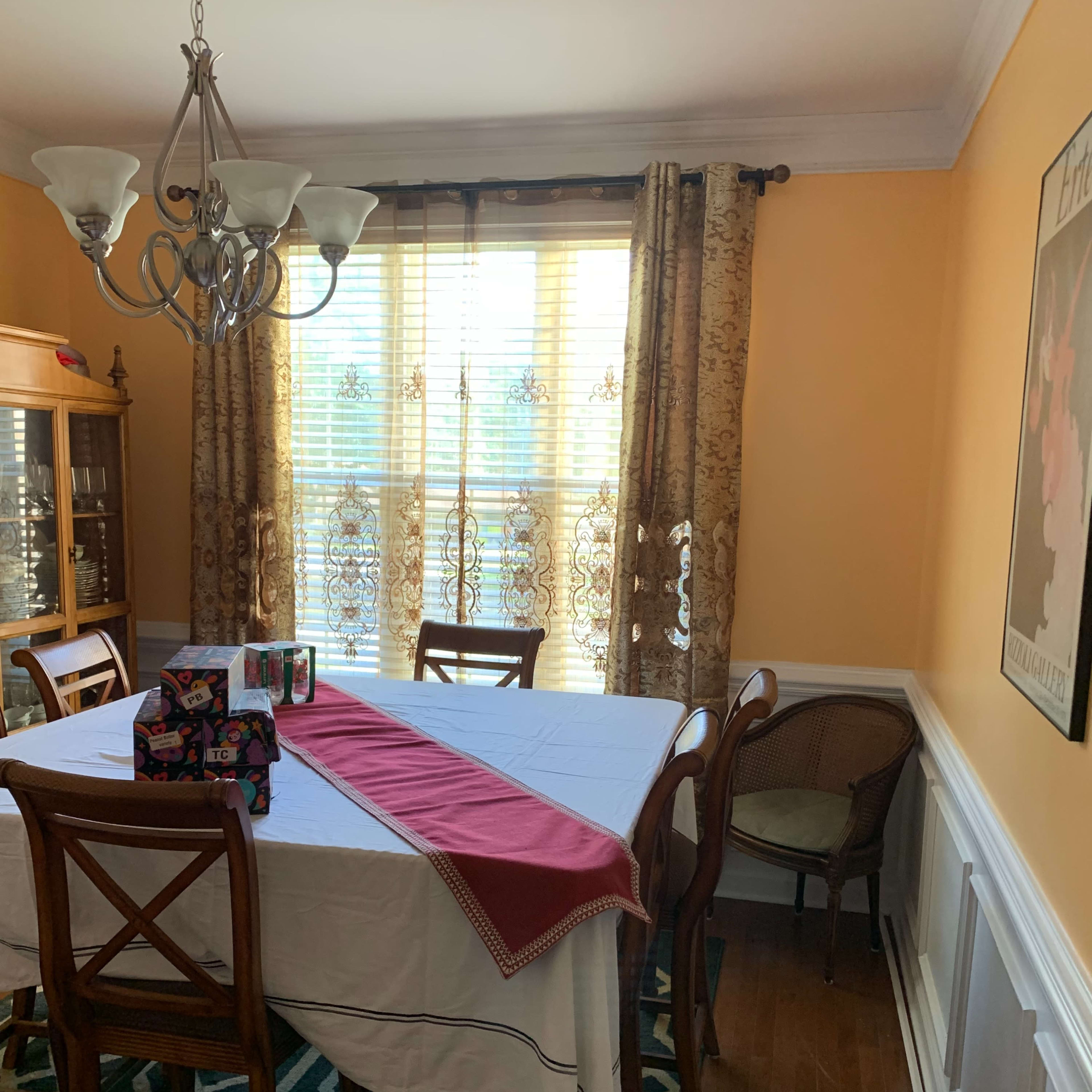 A dining room with a table covered by a white tablecloth, a red runner down the center, and several wrapped gifts on the table, surrounded by wooden chairs and a window with patterned curtains.