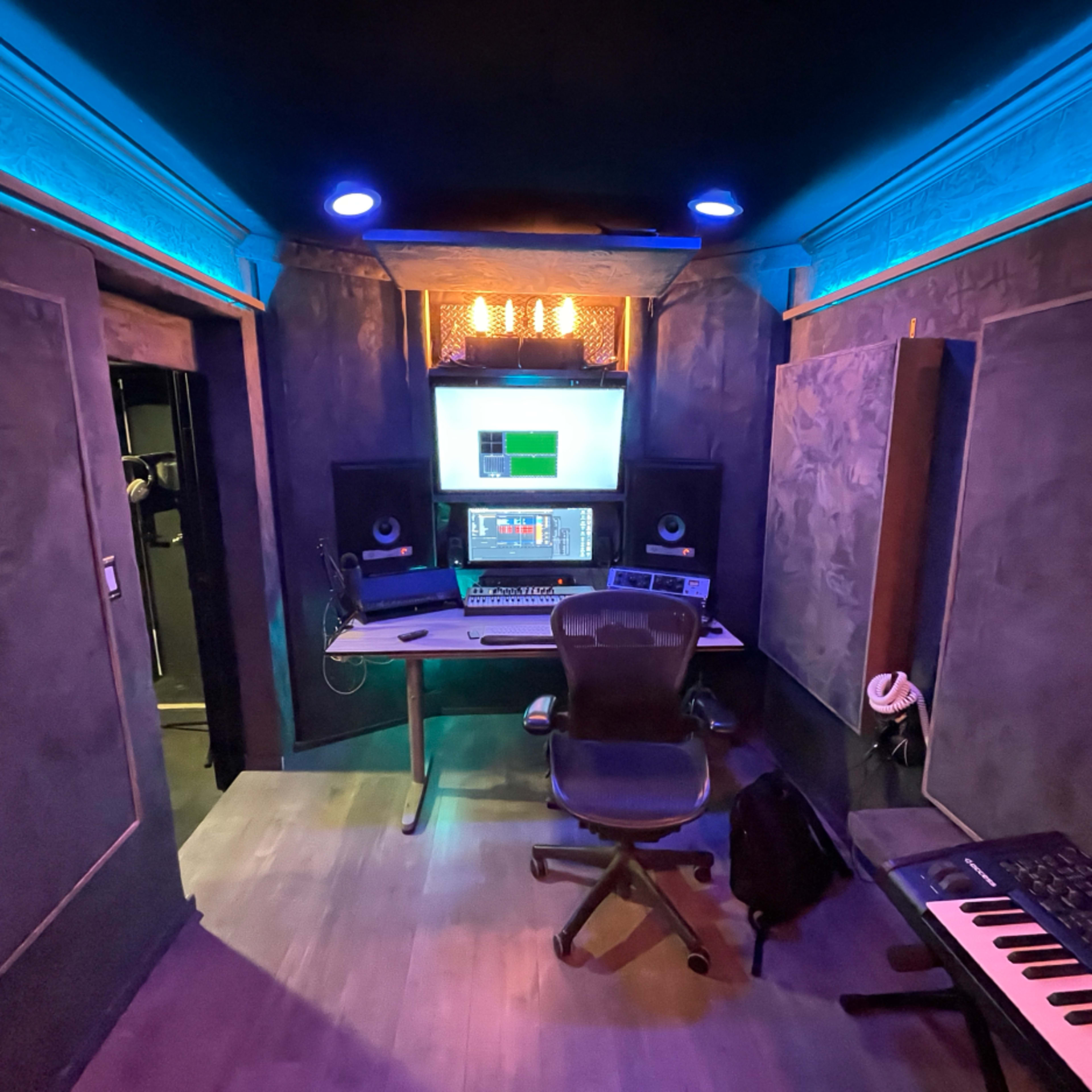 A music recording studio features a central workstation with a computer, audio equipment, and soundproof walls illuminated by blue and amber lights.