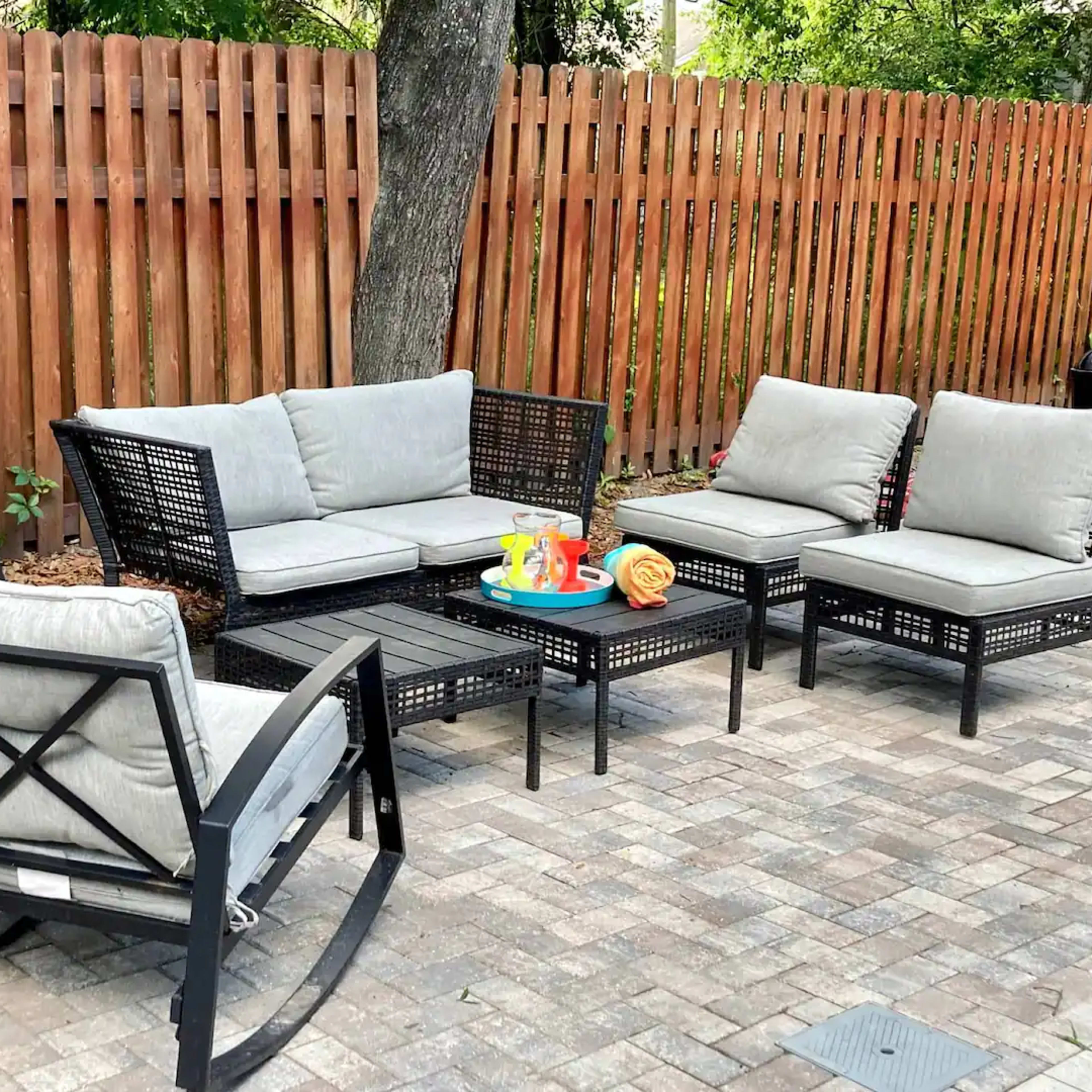 A backyard seating area with a black wicker furniture set, gray cushions, and a small coffee table, surrounded by a wooden fence and paved stones.
