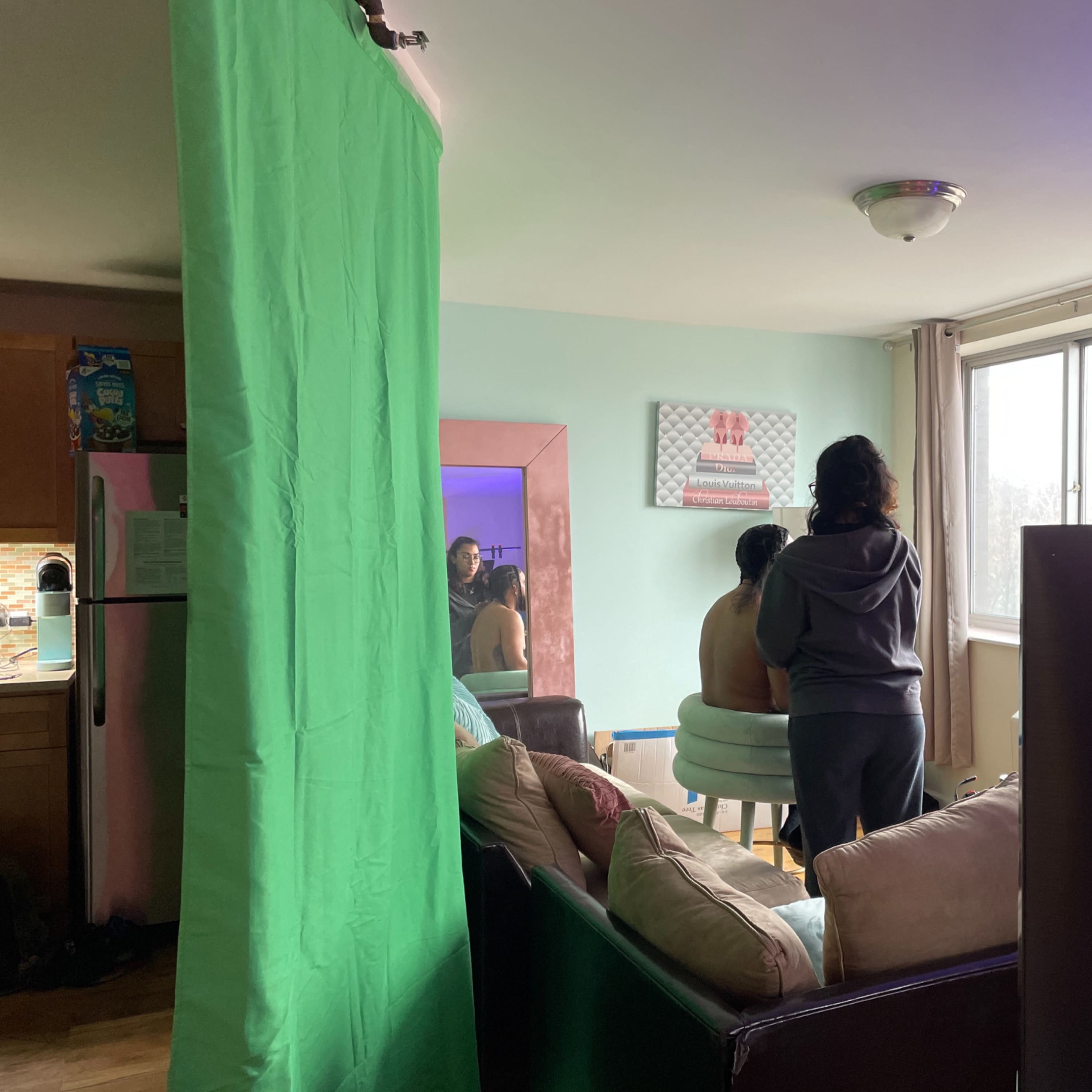 A person is seated in a chair while another person stands behind them, with a green screen and a television visible in a living room setting.
