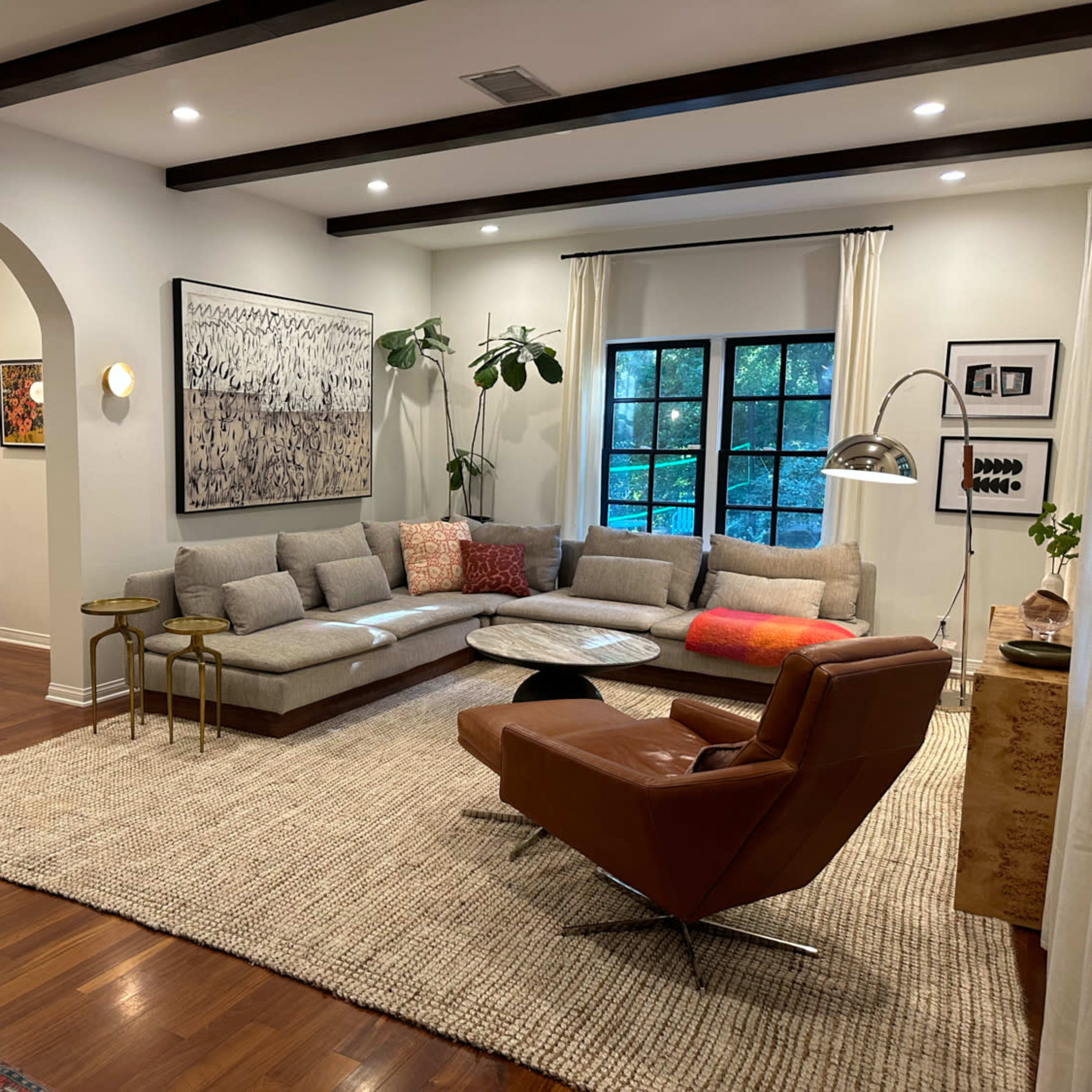 The living room features a sectional sofa, a round coffee table, and an accent chair, all arranged on a textured rug under softly lit walls with large windows.