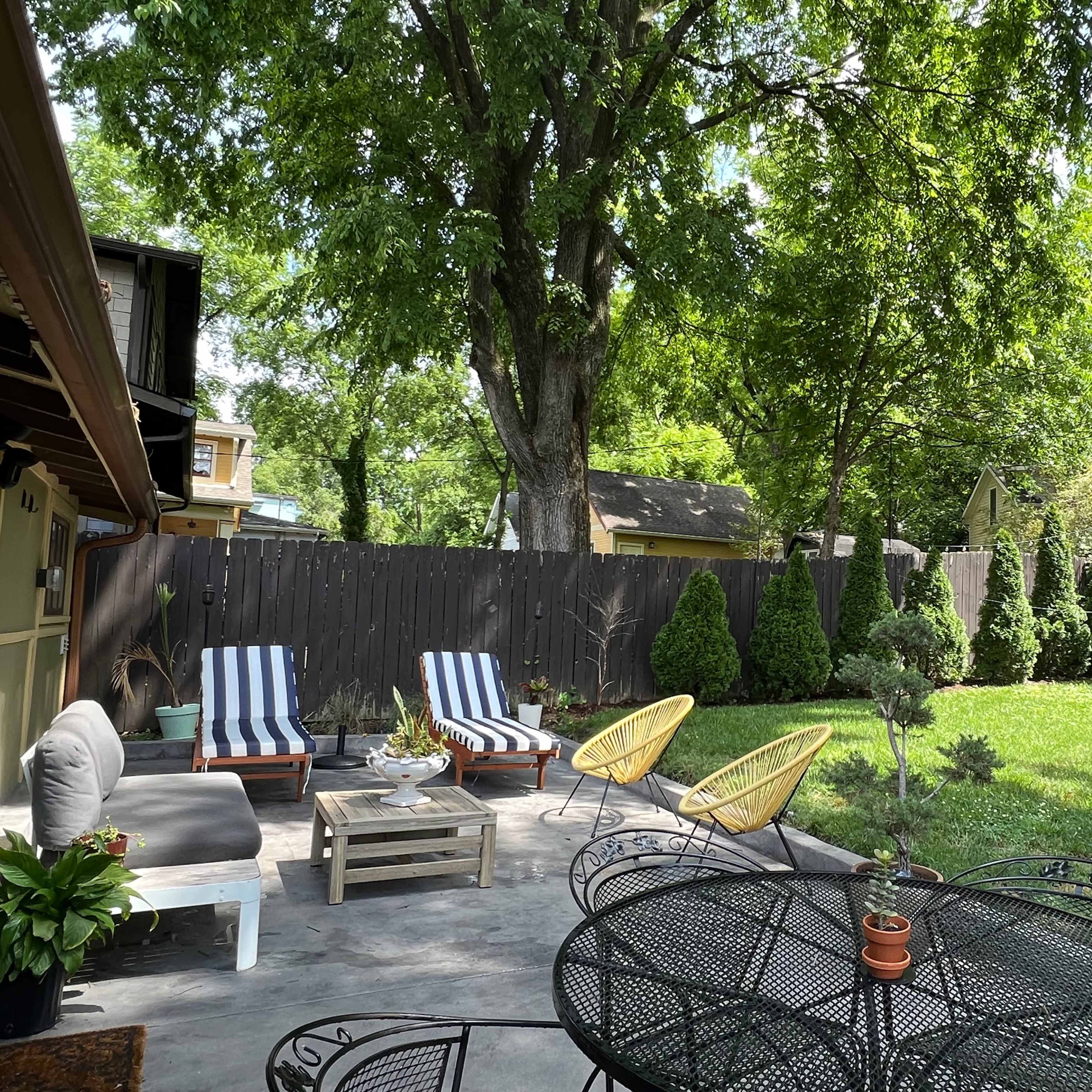 A patio area features outdoor seating with striped loungers, a round table, and a lush, green yard surrounded by trees and hedges.