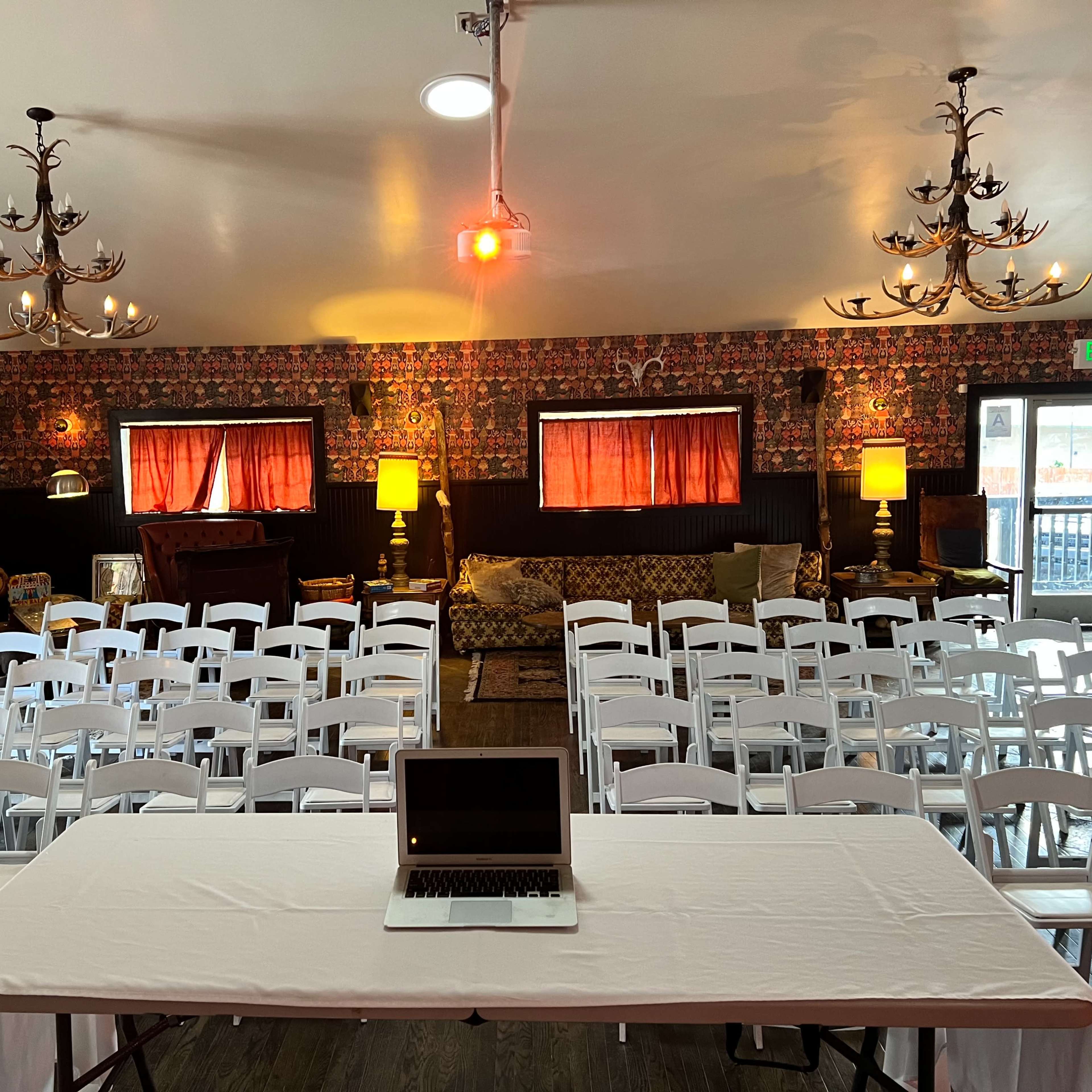 A room set up for an event, featuring rows of white chairs facing a table with a laptop, and decorated with patterned wallpaper and chandeliers.