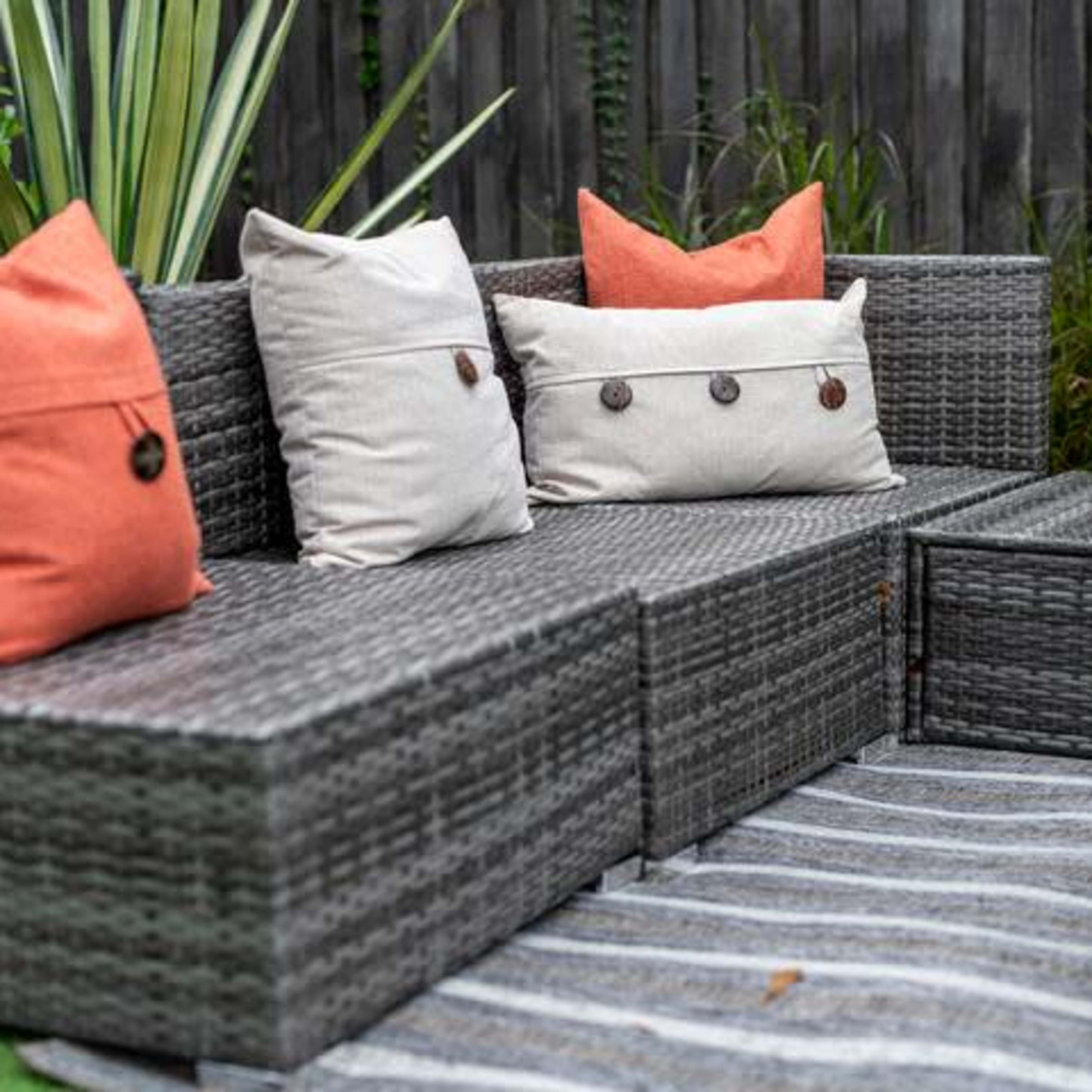 A sectional outdoor couch made of wicker, with beige and orange pillows, positioned on a striped rug in a garden setting.