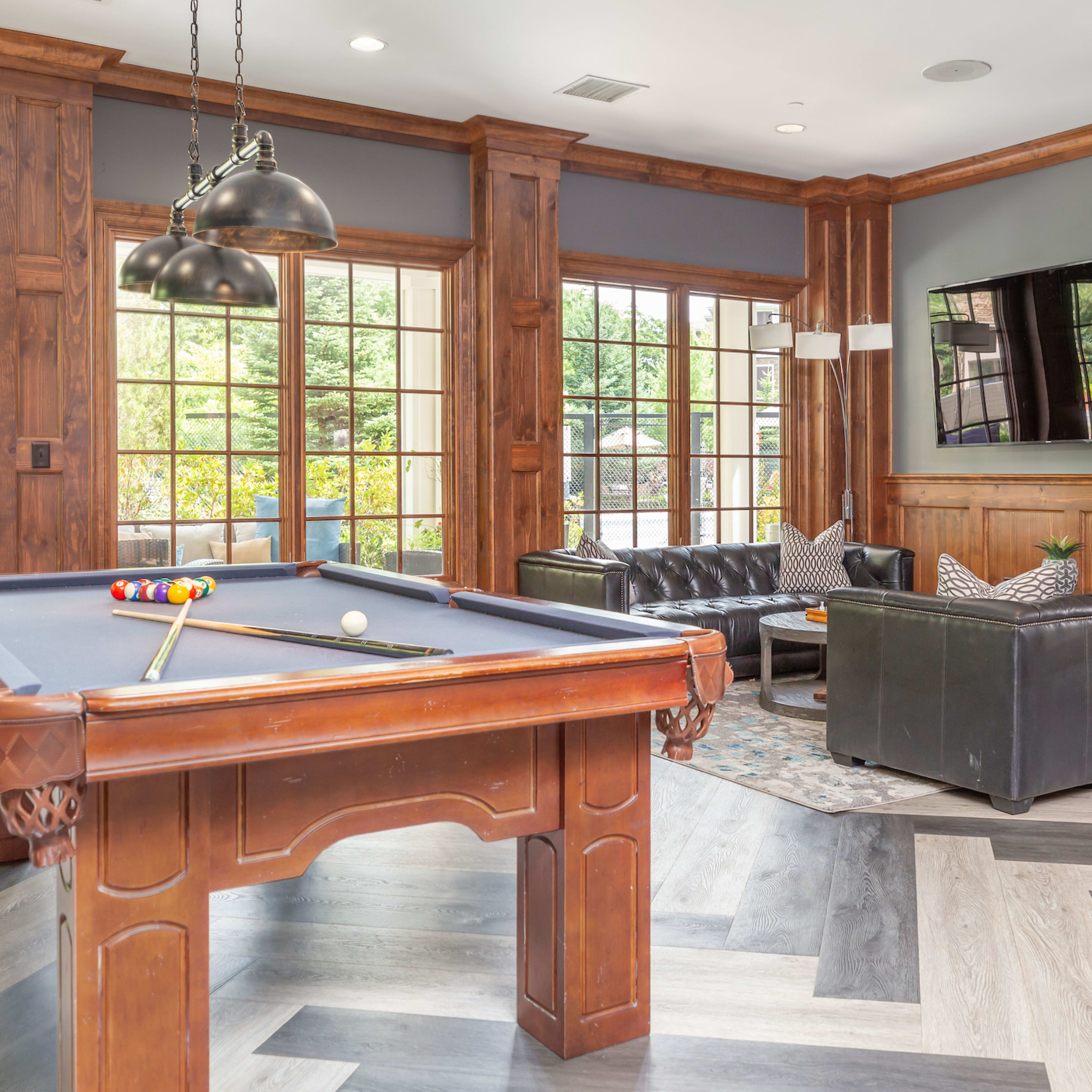 A billiard table in a lounge area with wooden paneling, comfortable seating, and large windows allowing natural light.