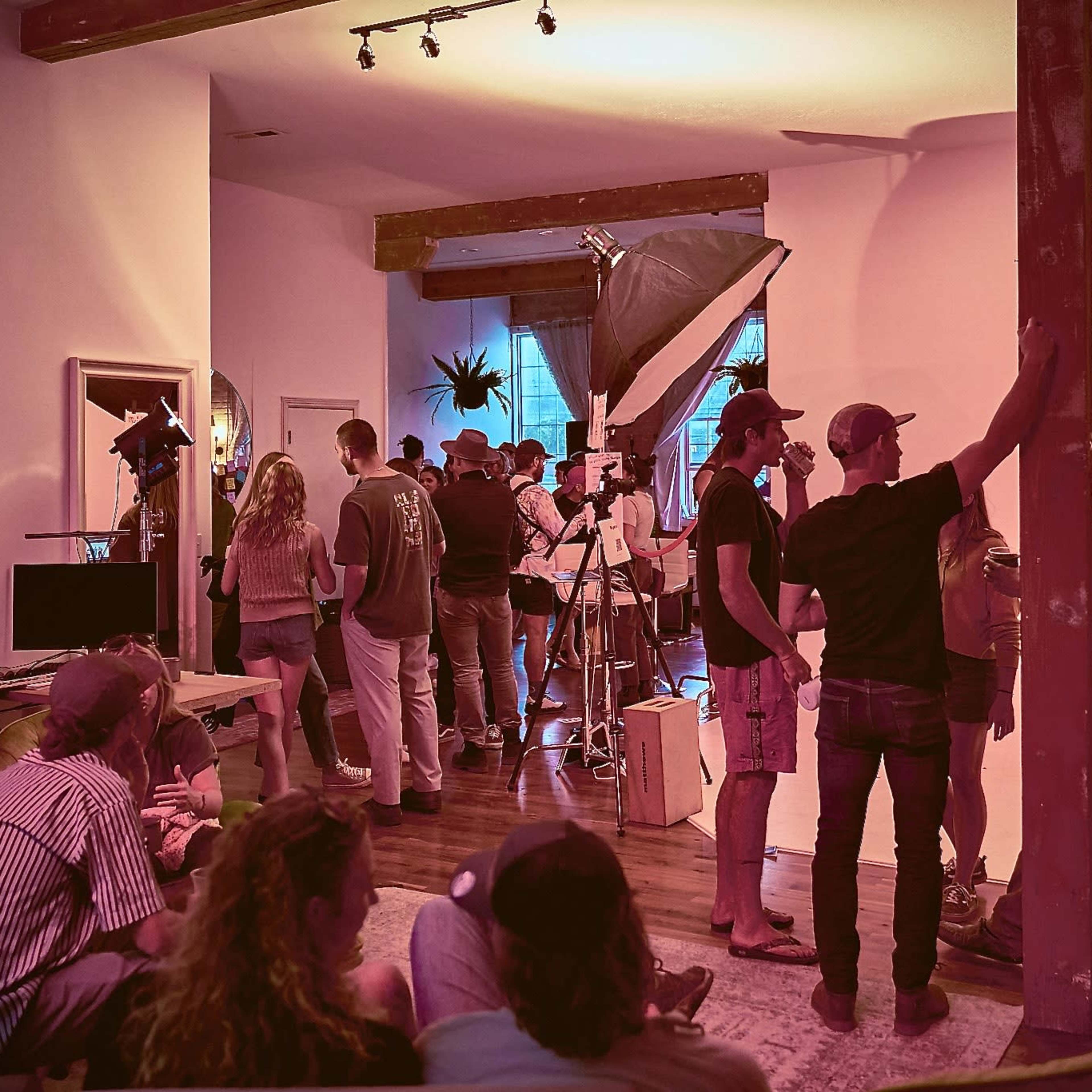 A group of people gathers in a room with studio equipment and soft lighting, while some are engaged in conversation and others are preparing for a shoot.