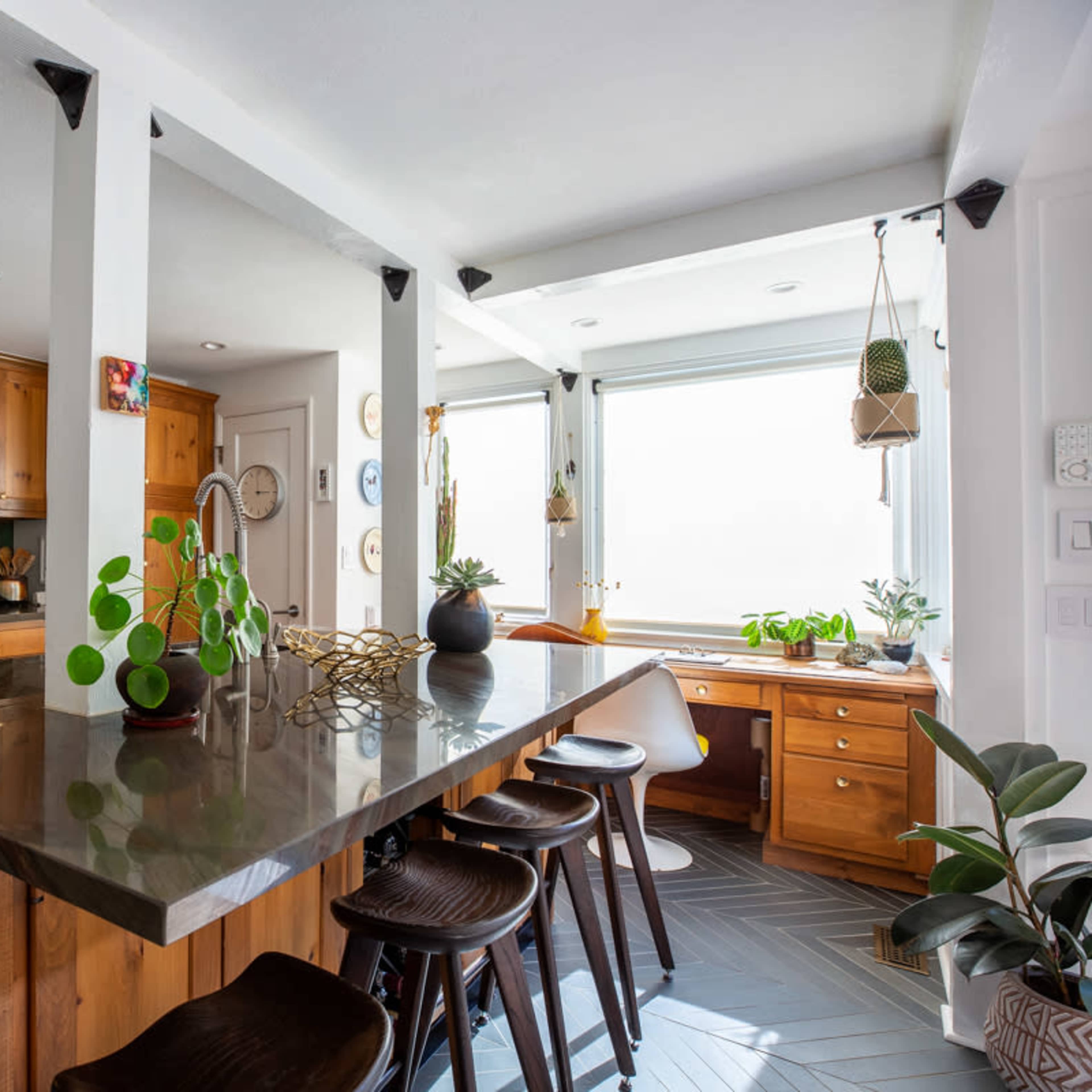 A kitchen with wooden cabinets, a large island with dark countertops, and a small workspace by a window, featuring plants and natural light.