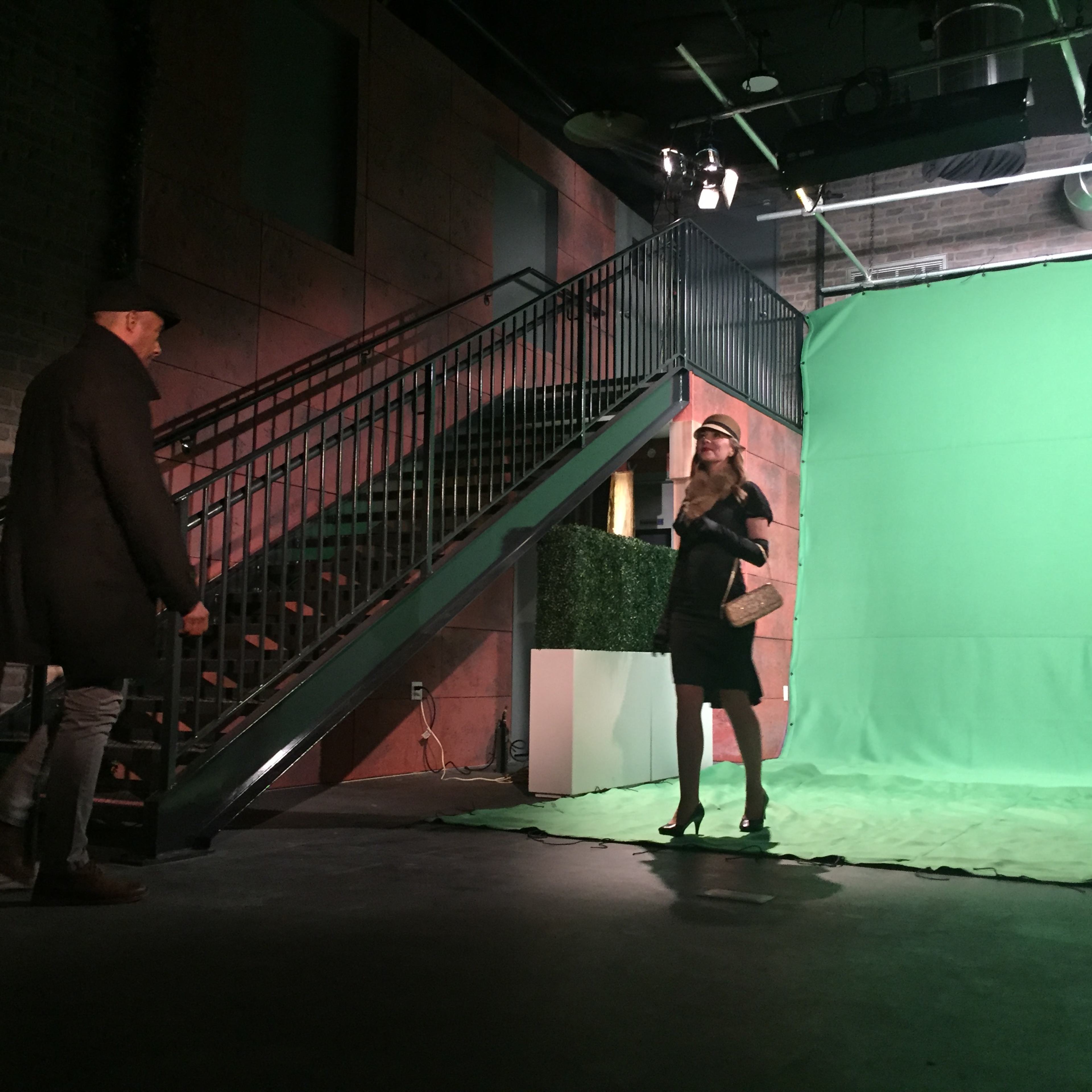 A man stands at the bottom of a staircase while a woman poses on a green screen stage.