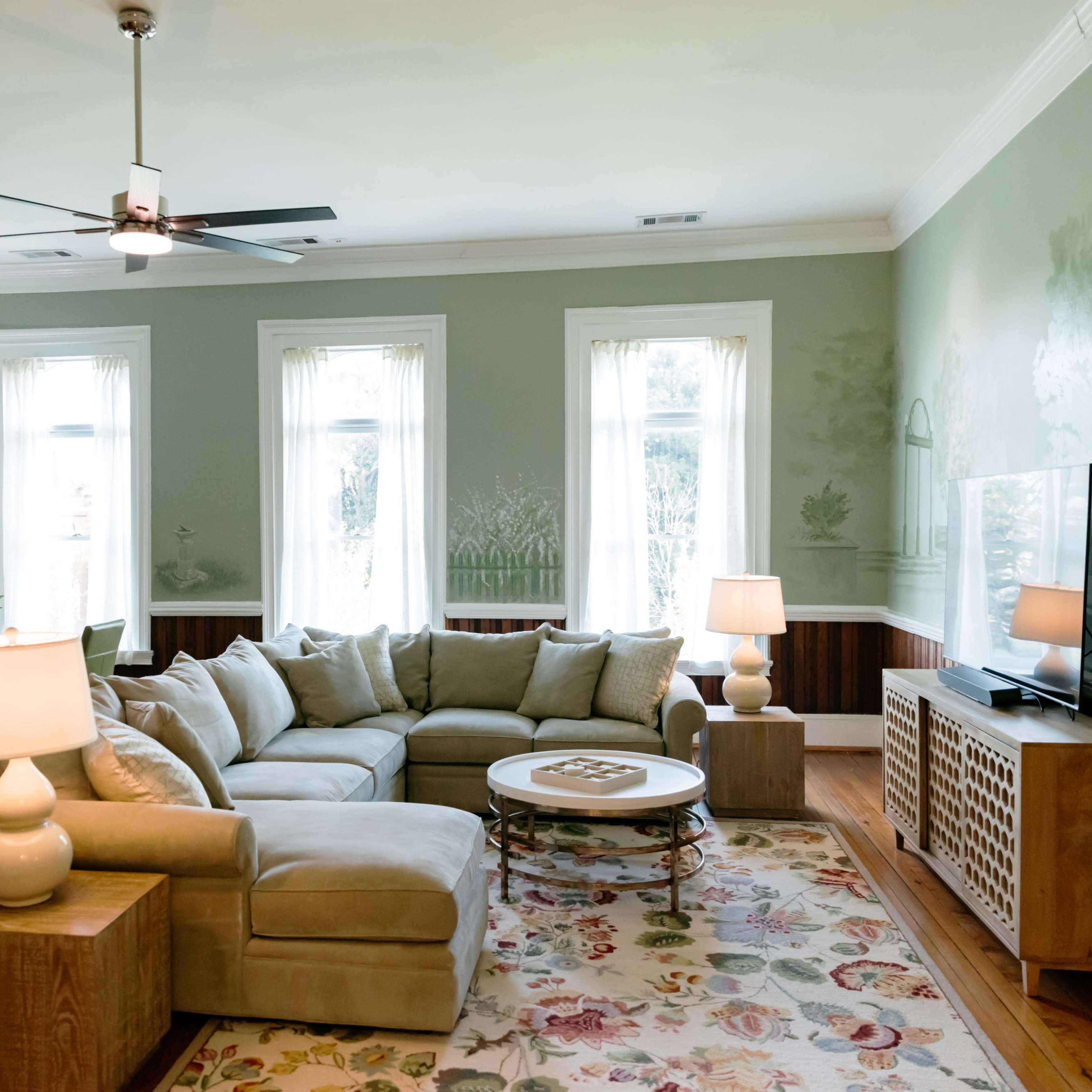A spacious living room features a light-colored sectional sofa, a coffee table, a television, and large windows allowing natural light, with a dining area visible in the background.