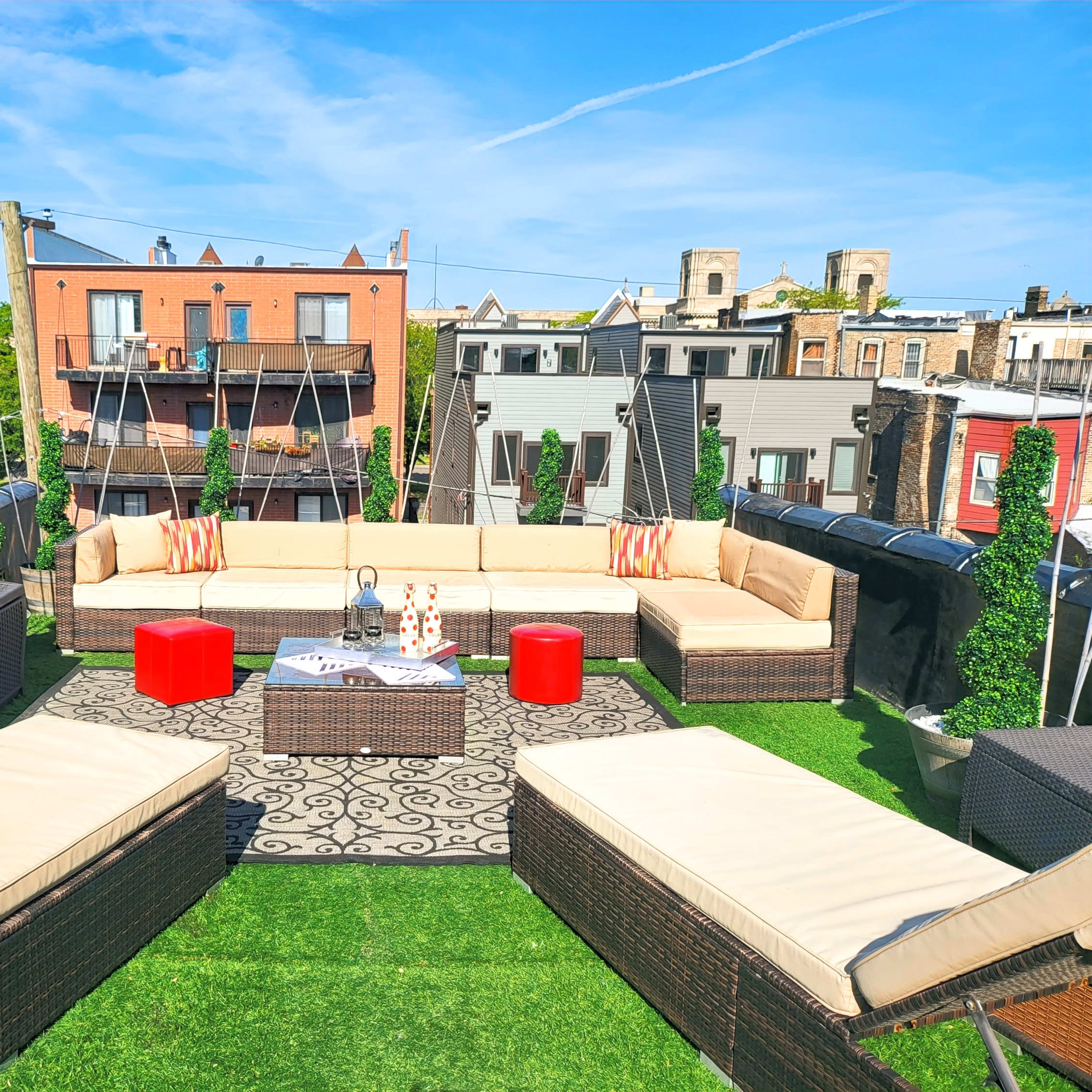 A rooftop terrace features an arrangement of sectional couches, red stools, and potted greenery under a clear blue sky.