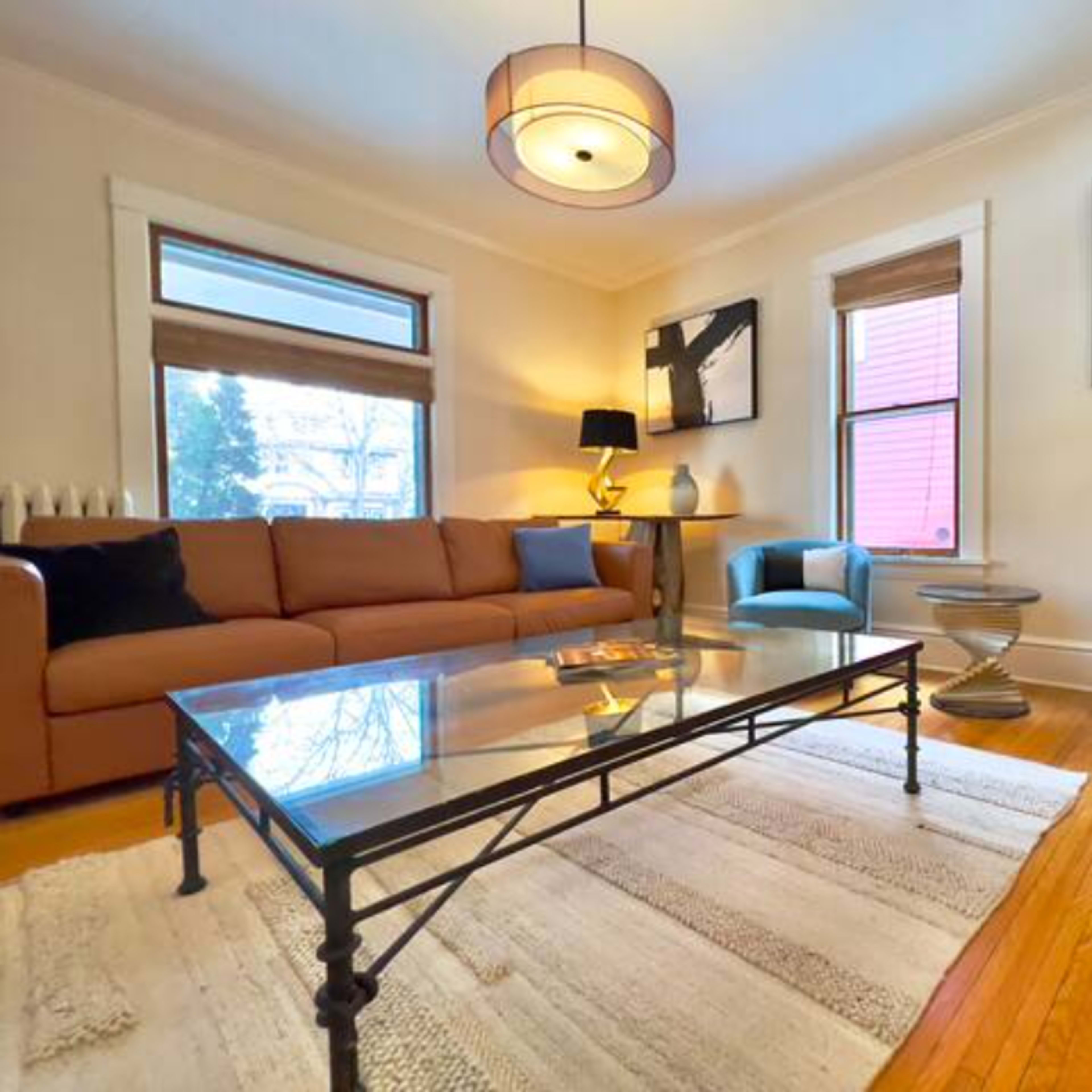 A living room with a brown sofa, two blue chairs, a glass coffee table, and large windows letting in natural light.