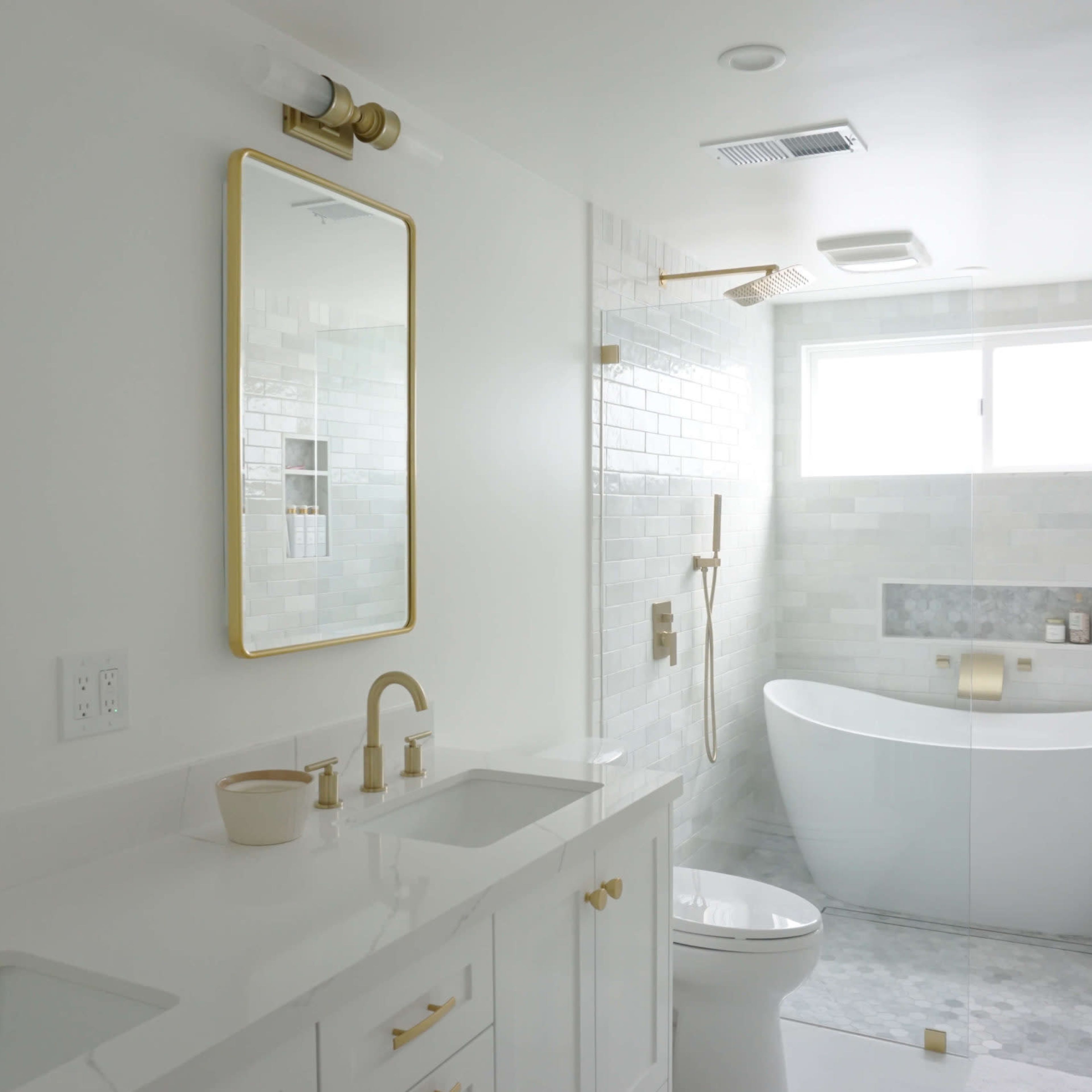 A modern bathroom features dual mirrors above a white countertop with gold fixtures, a freestanding tub, and a large window providing natural light.