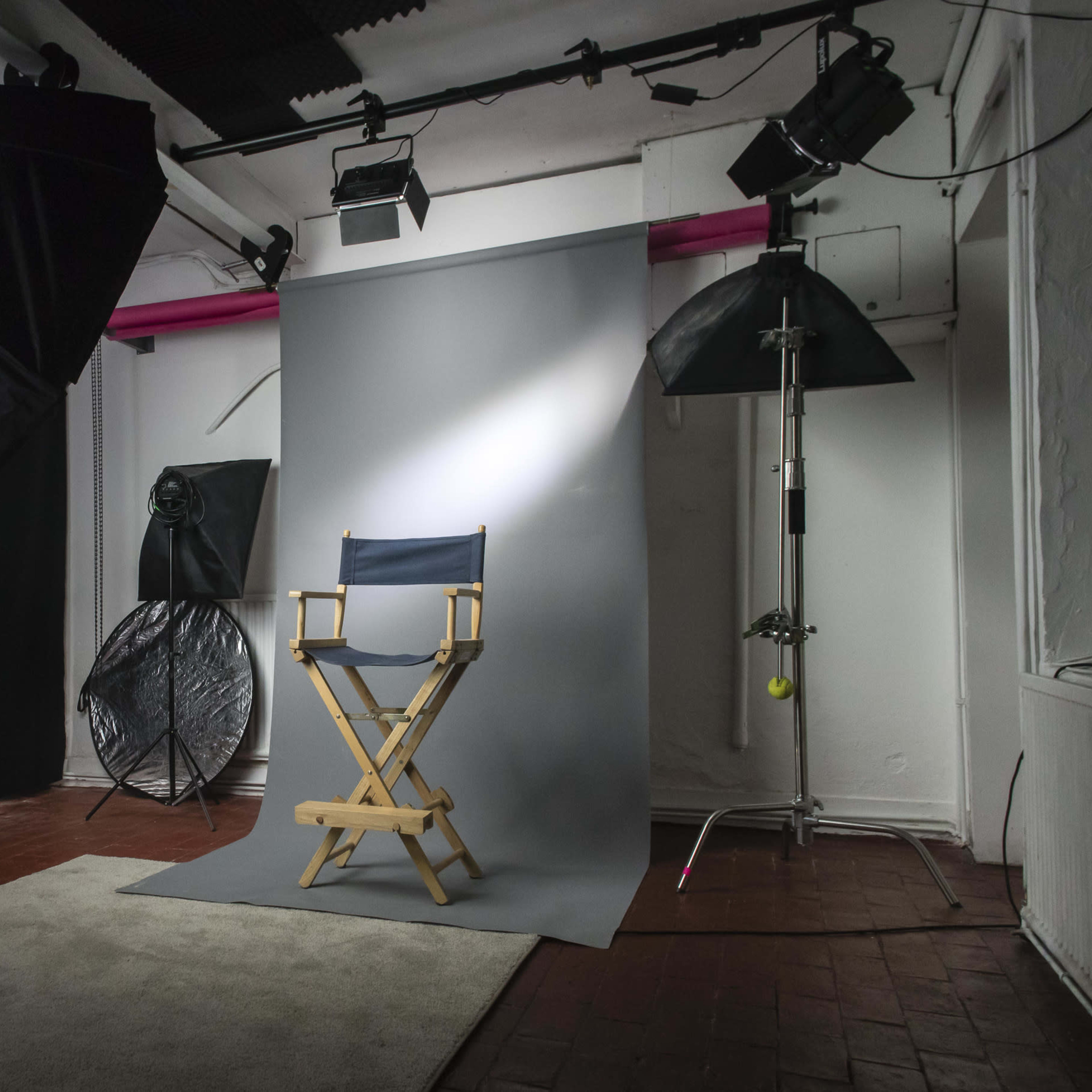 A photography studio setup with a director's chair positioned in front of a gray backdrop, surrounded by various lighting equipment and a carpeted area.