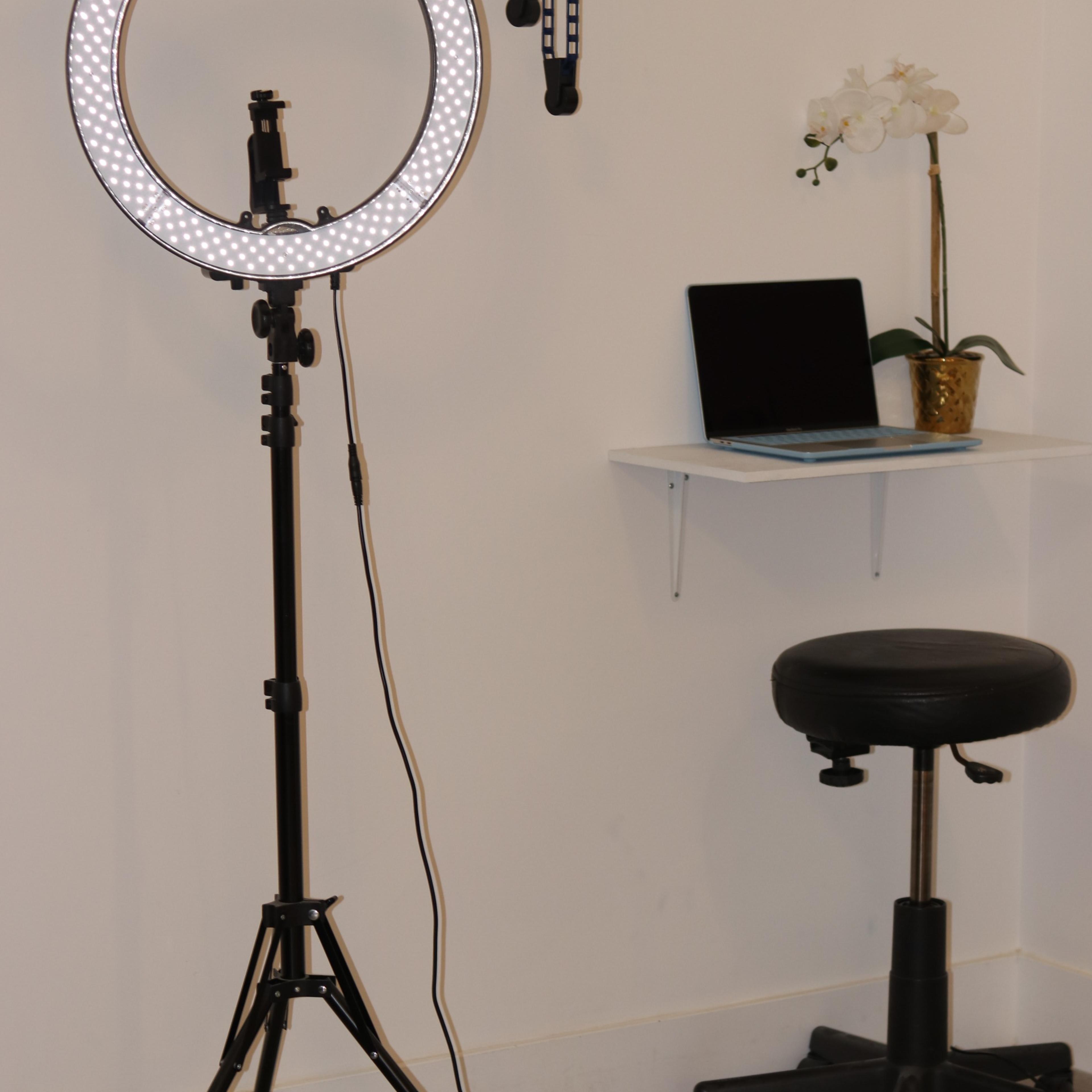 A ring light is mounted on a tripod next to a small desk with a laptop and a potted plant, along with a black rolling stool.