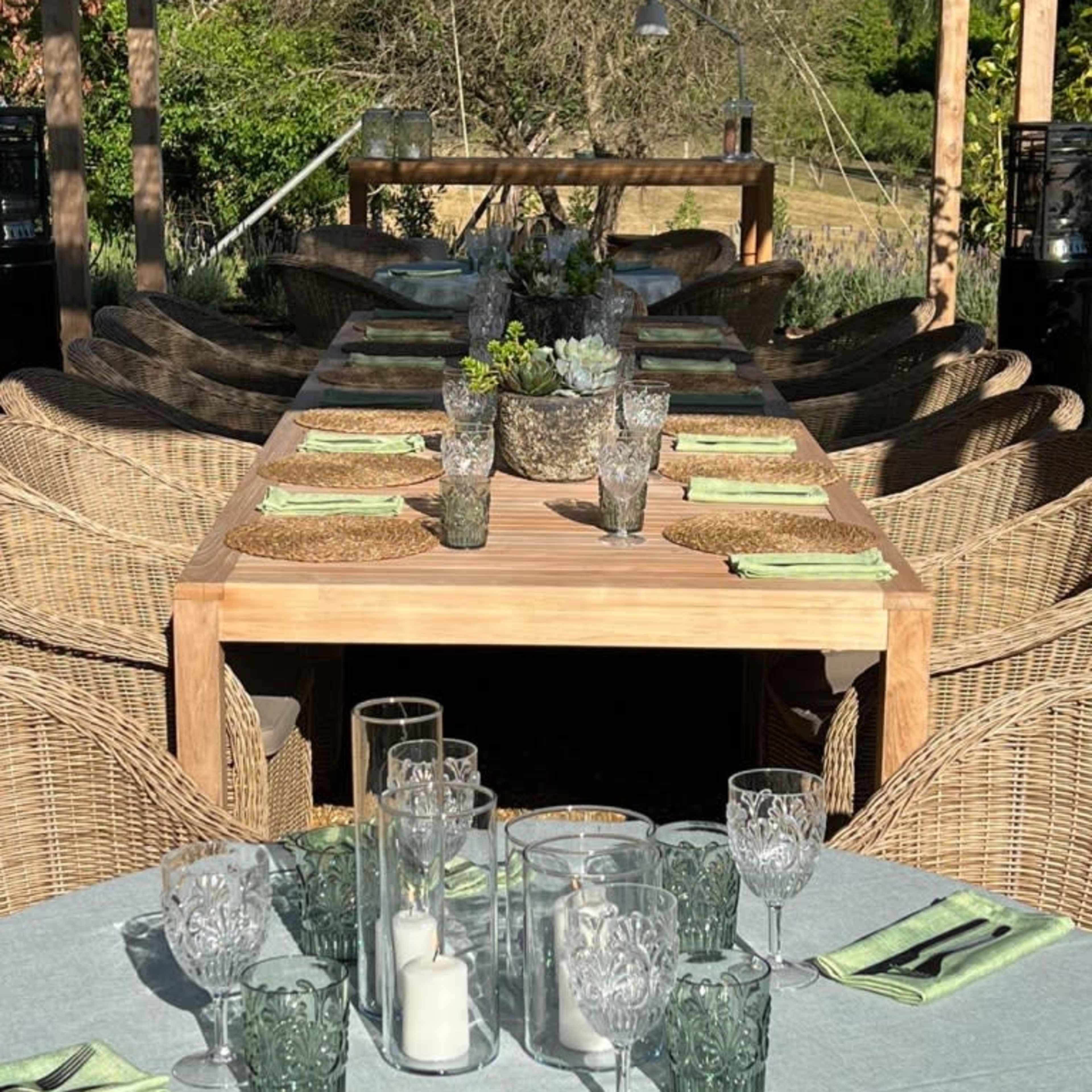 A long wooden table is set for a meal, surrounded by wicker chairs, with decorative glassware and place settings arranged neatly.