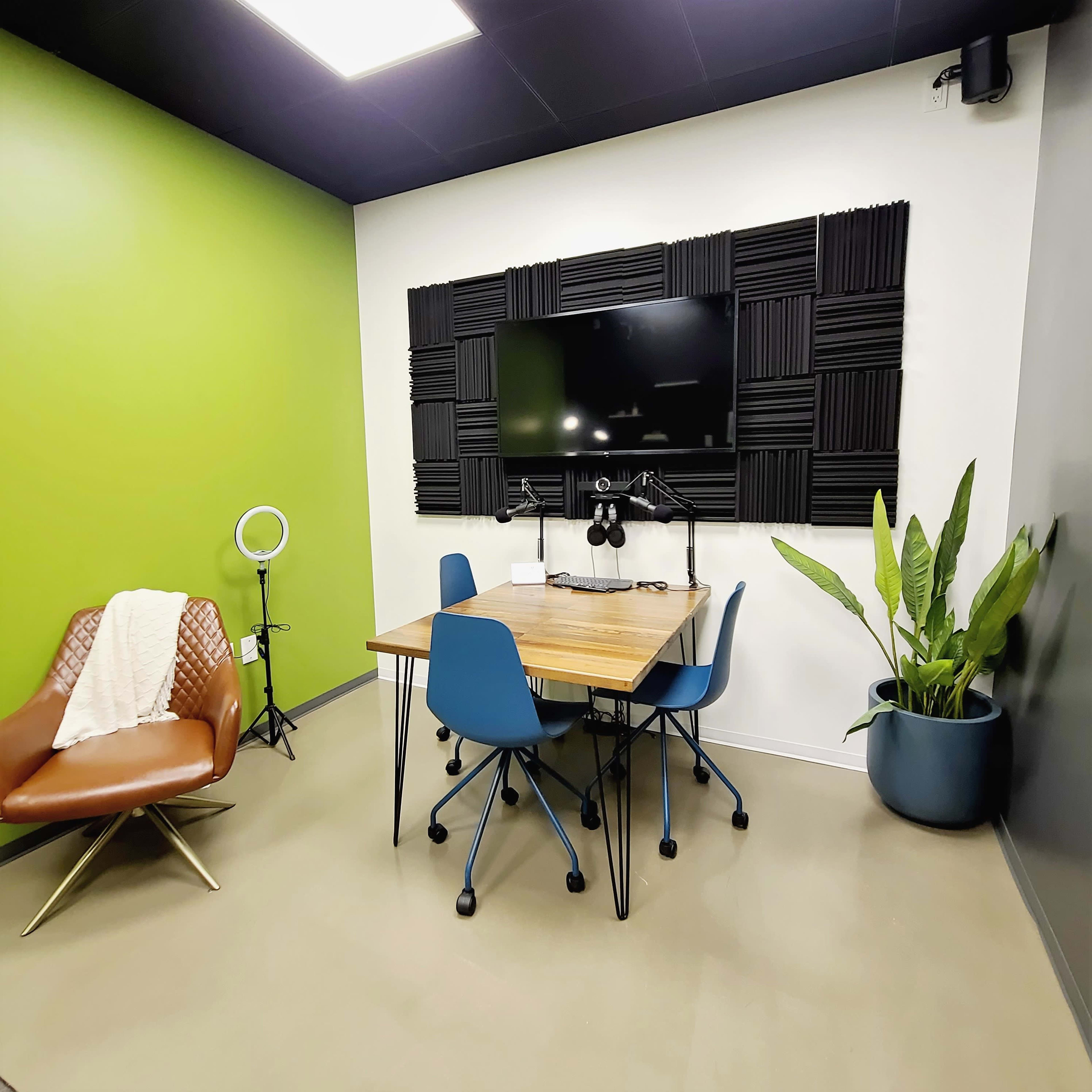 A small meeting room with a wooden table surrounded by four blue chairs, a wall-mounted TV, acoustic panels, a green accent wall, a plant in a pot, and a ring light next to a brown leather chair.