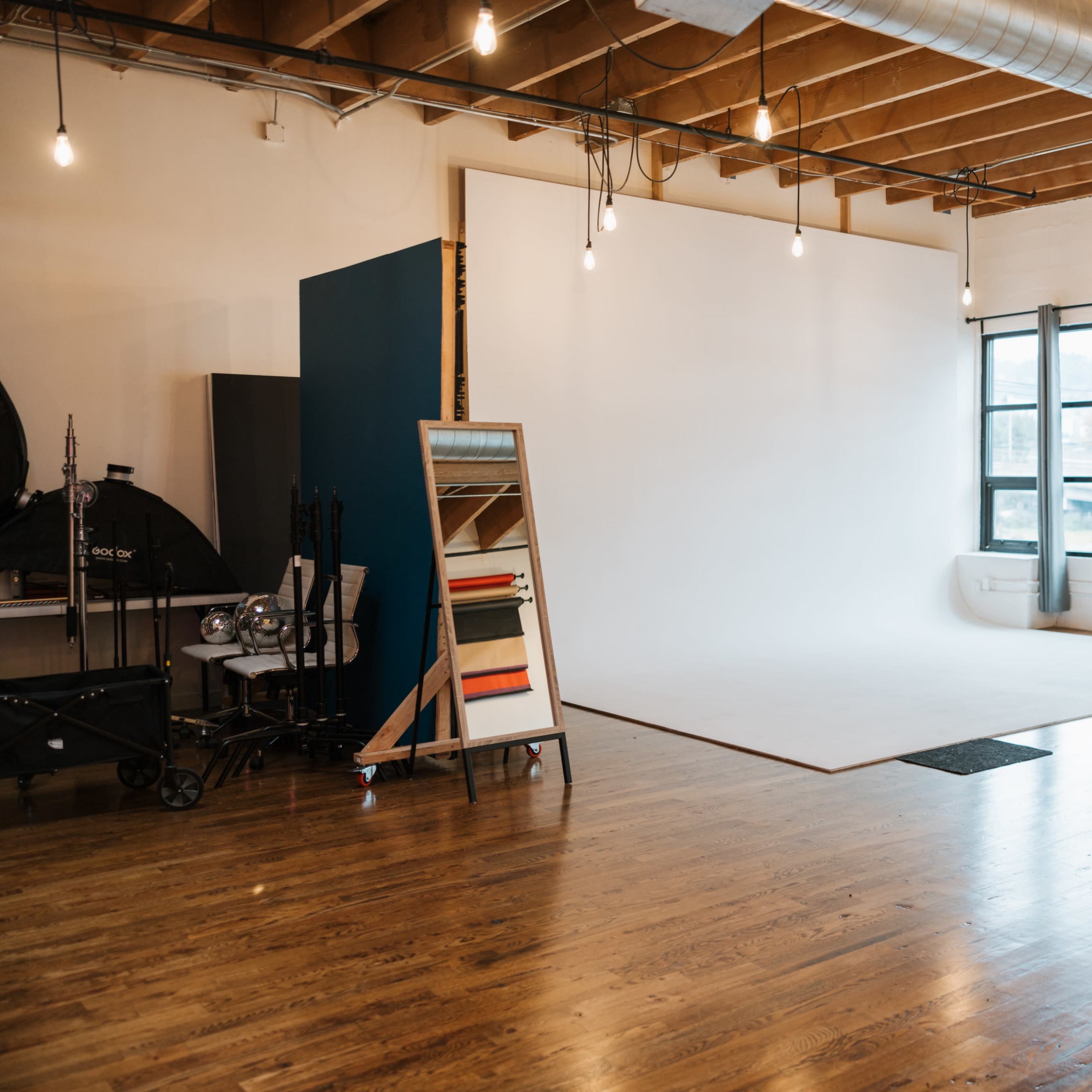 A spacious photo studio with a plain white backdrop, large windows, and wooden flooring, featuring a mirror and various equipment along one side.