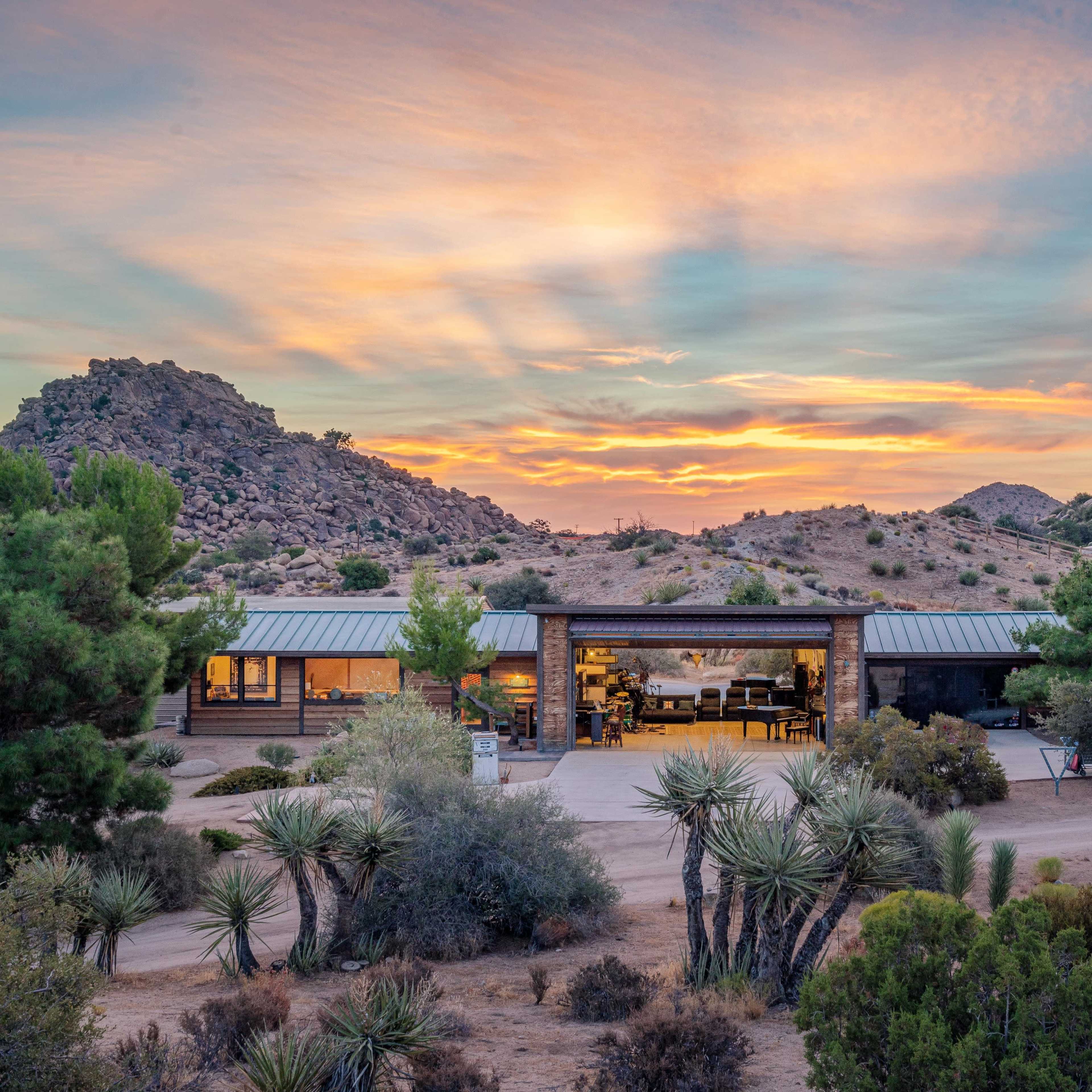 A modern house set against a backdrop of mountains and a colorful sunset, surrounded by desert vegetation.