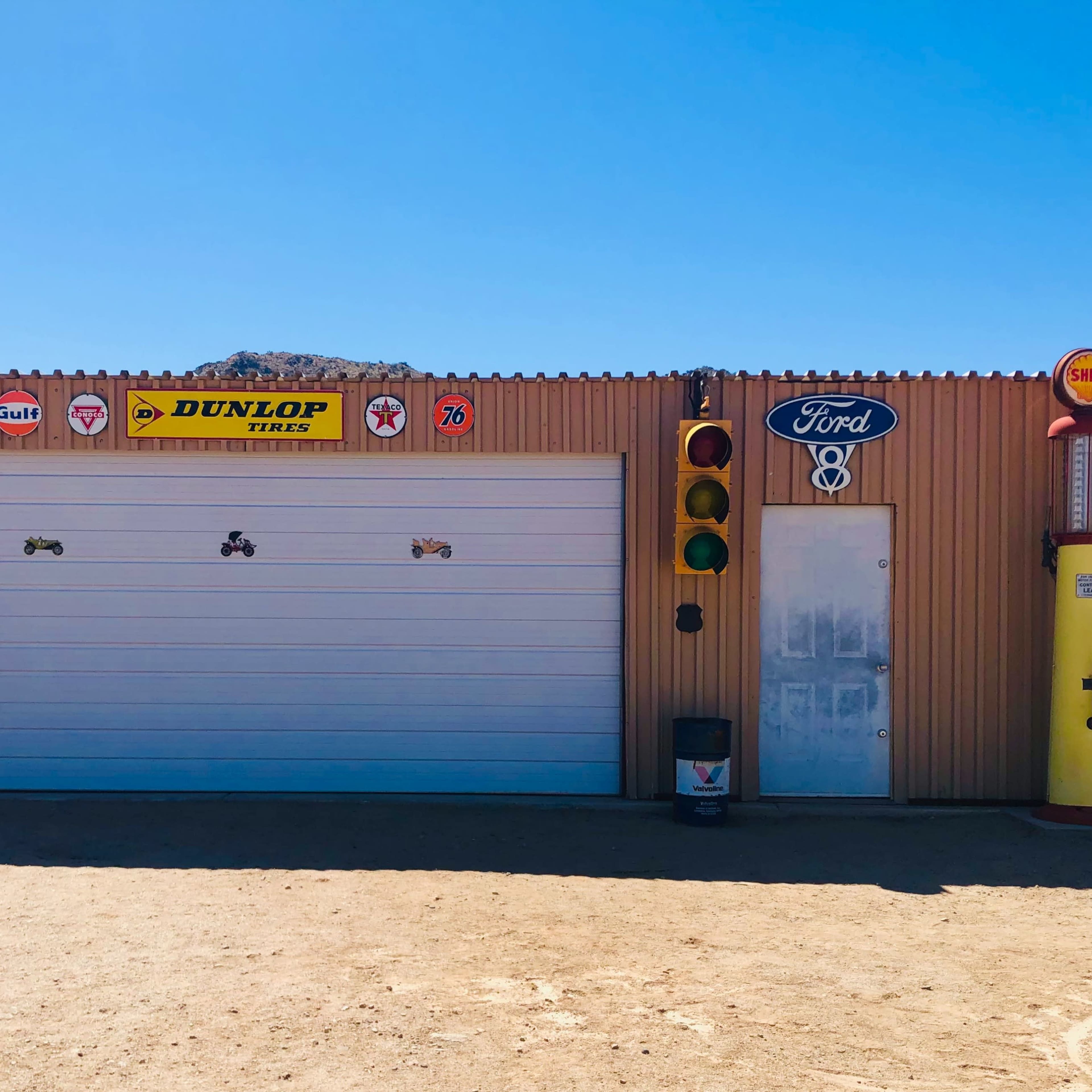 A metal building with two closed garage doors displays vintage gas station signs and memorabilia, including a Ford sign and a traffic light, in a rural setting.