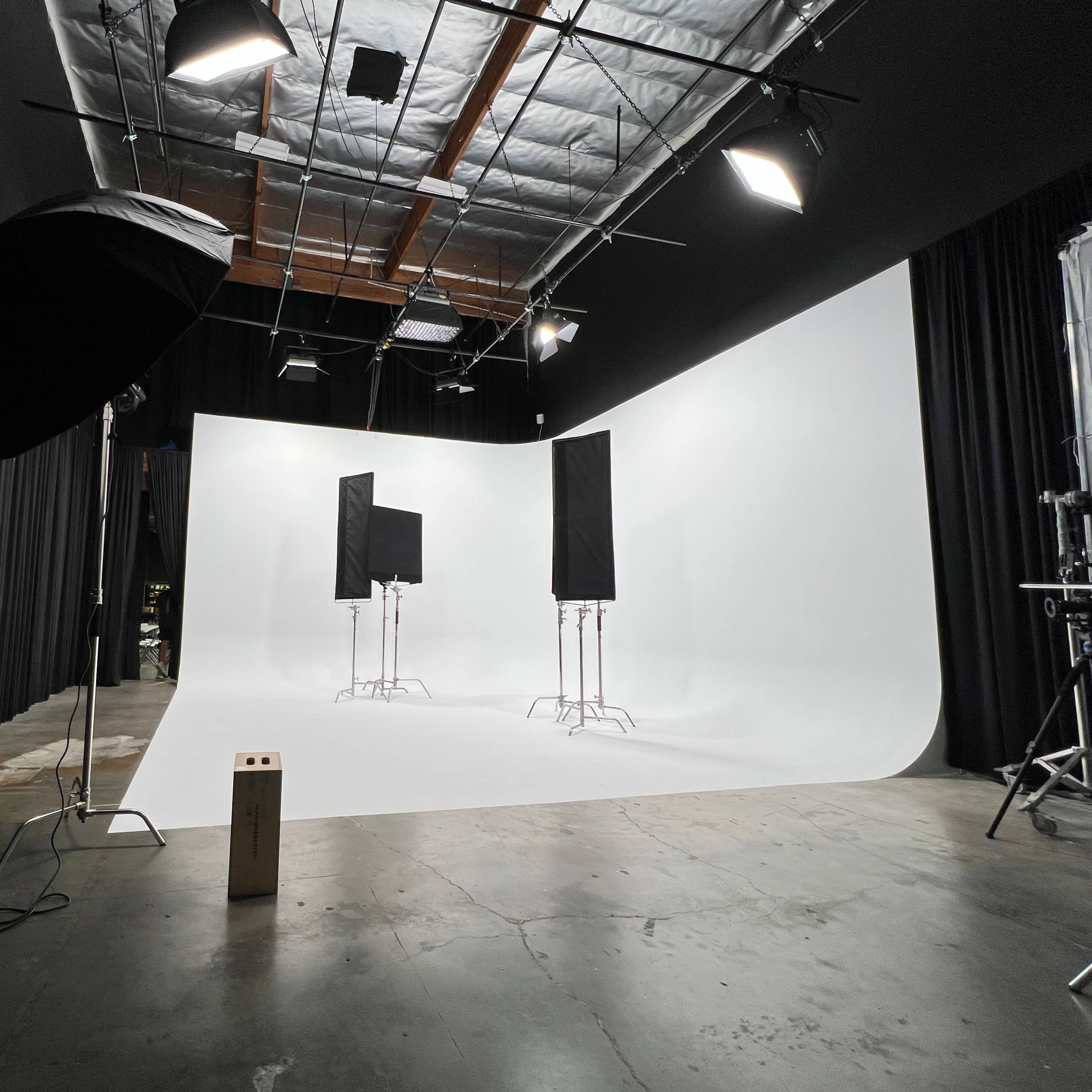 A photography studio with a white backdrop, three light stands with softboxes, and a dark curtain lining the walls.