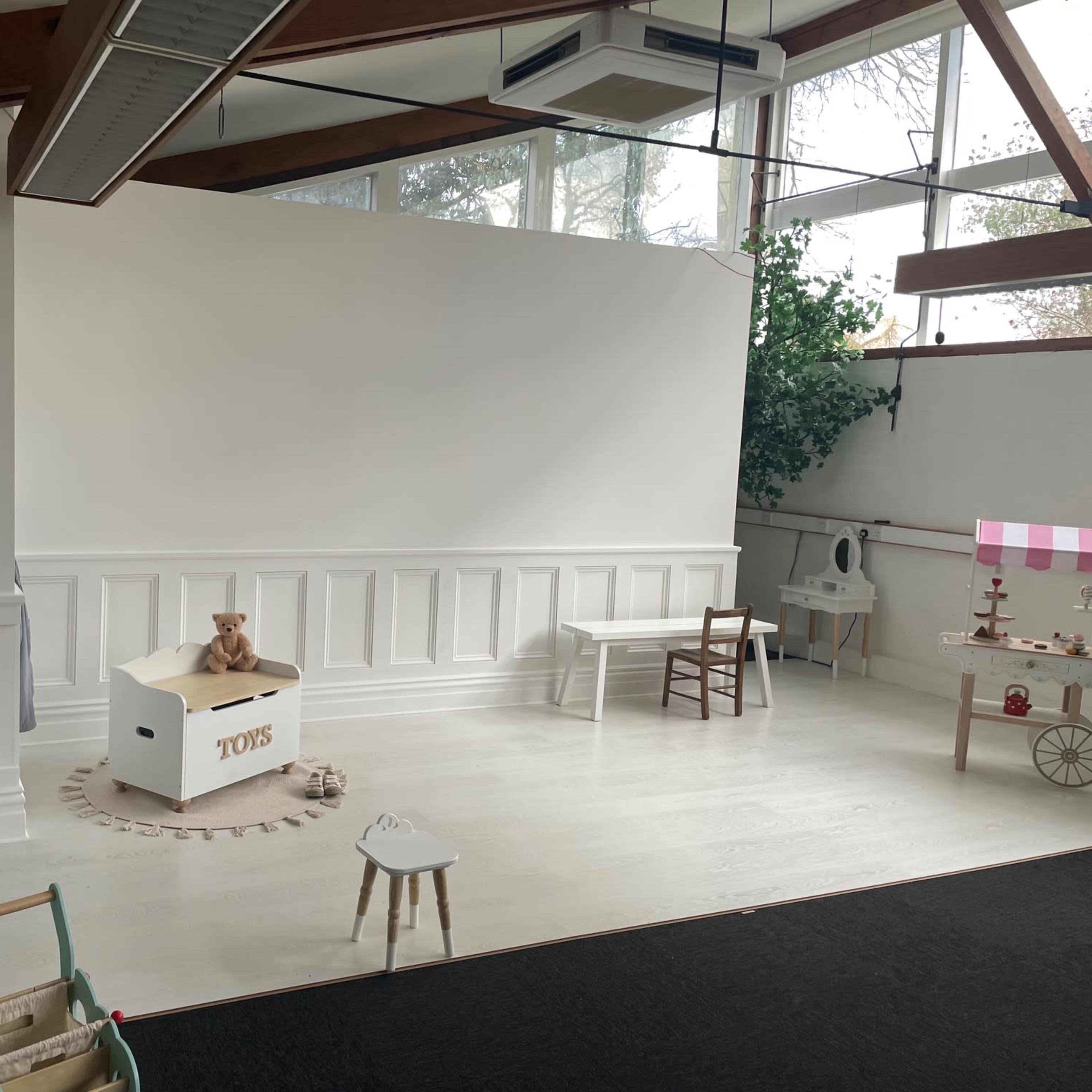 A spacious room with a white wall, wooden beams, and minimal furniture, including a toy box, a small table, and a chair.