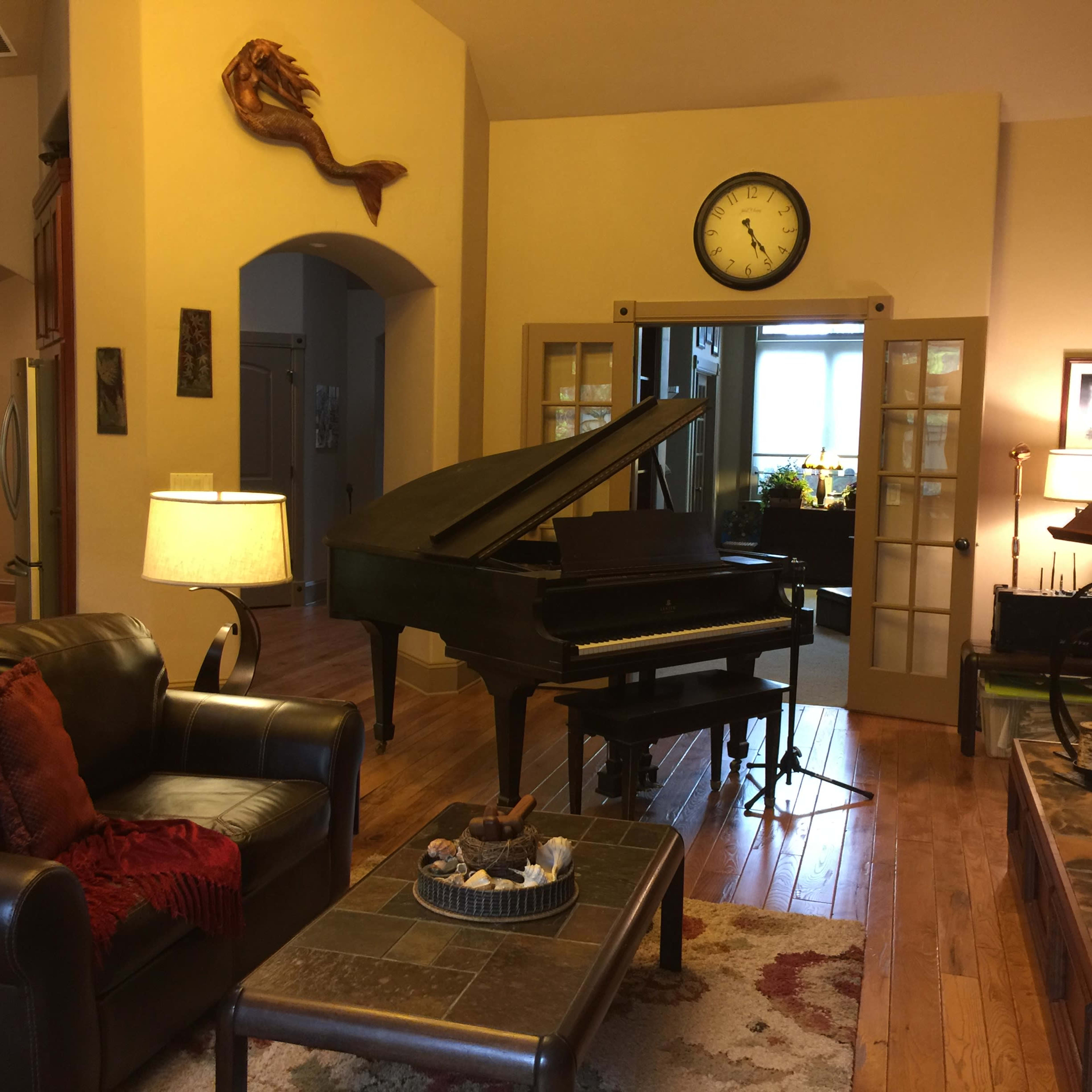 A cozy living space featuring a grand piano, a leather sofa with red throw pillows, and a stone fireplace, complemented by warm lighting and wooden floors.