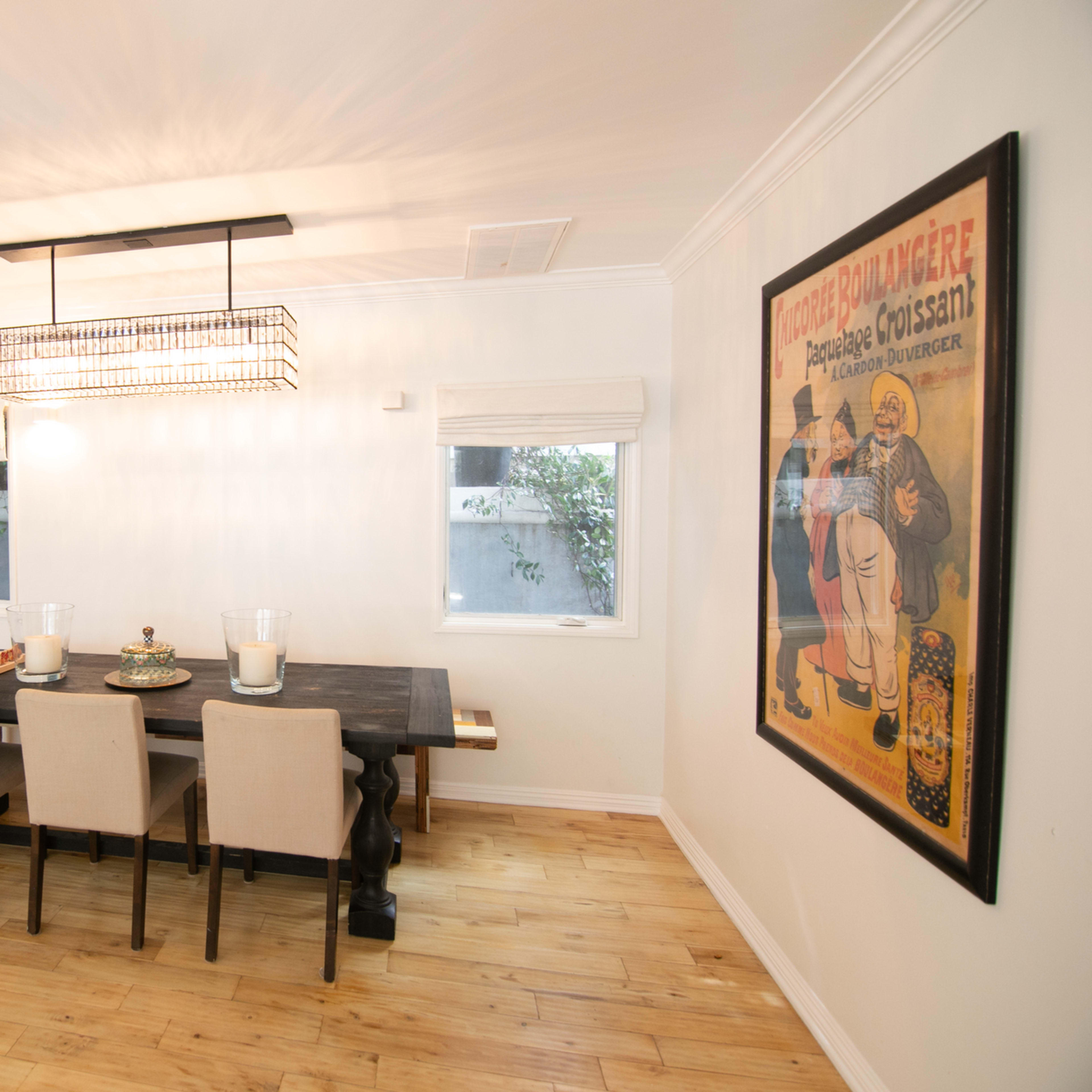 A dining area features a long table with four beige chairs, illuminated by a modern light fixture, and two framed posters on the walls.