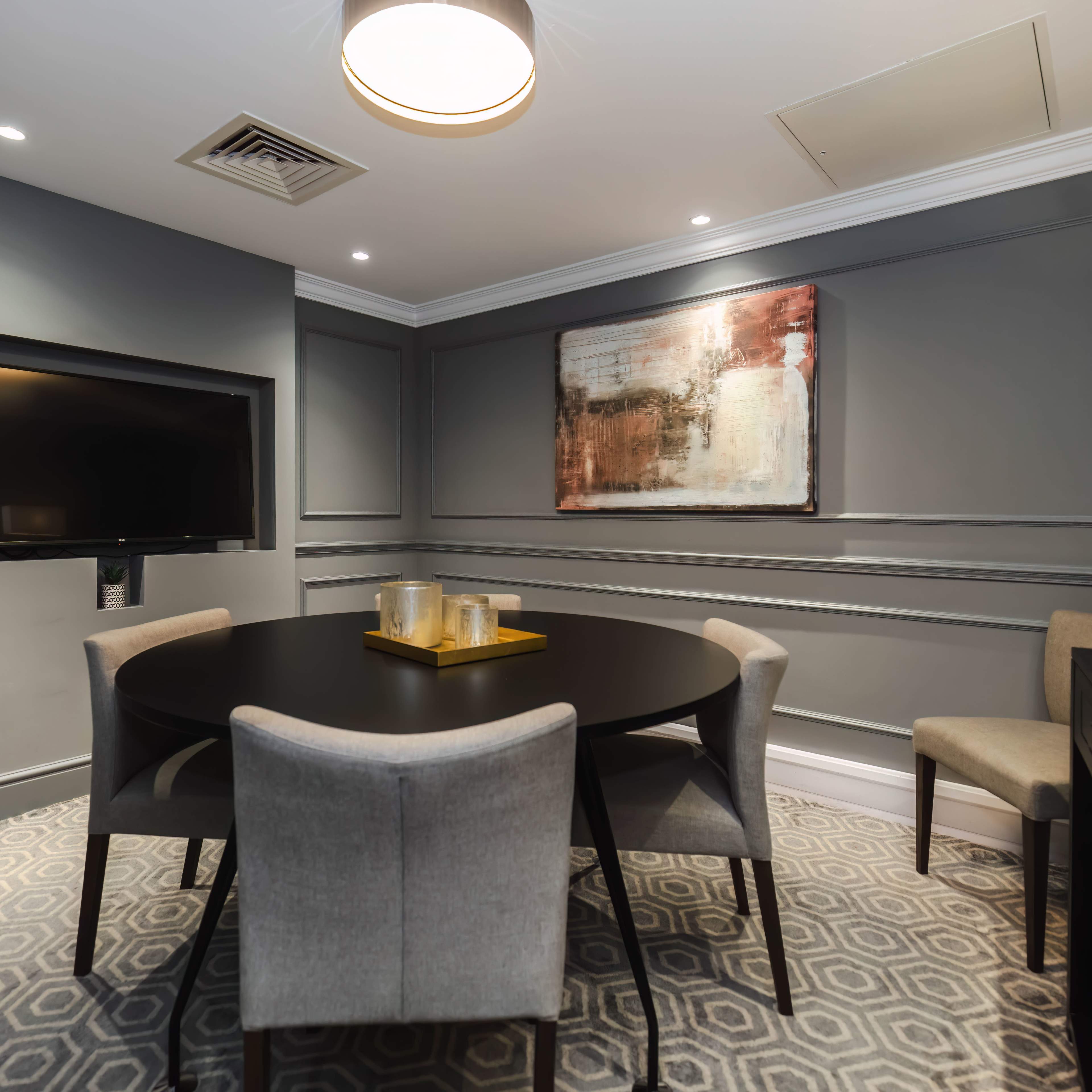 A small dining area with a round black table surrounded by four grey chairs, a wall-mounted television, and an abstract painting on the wall.