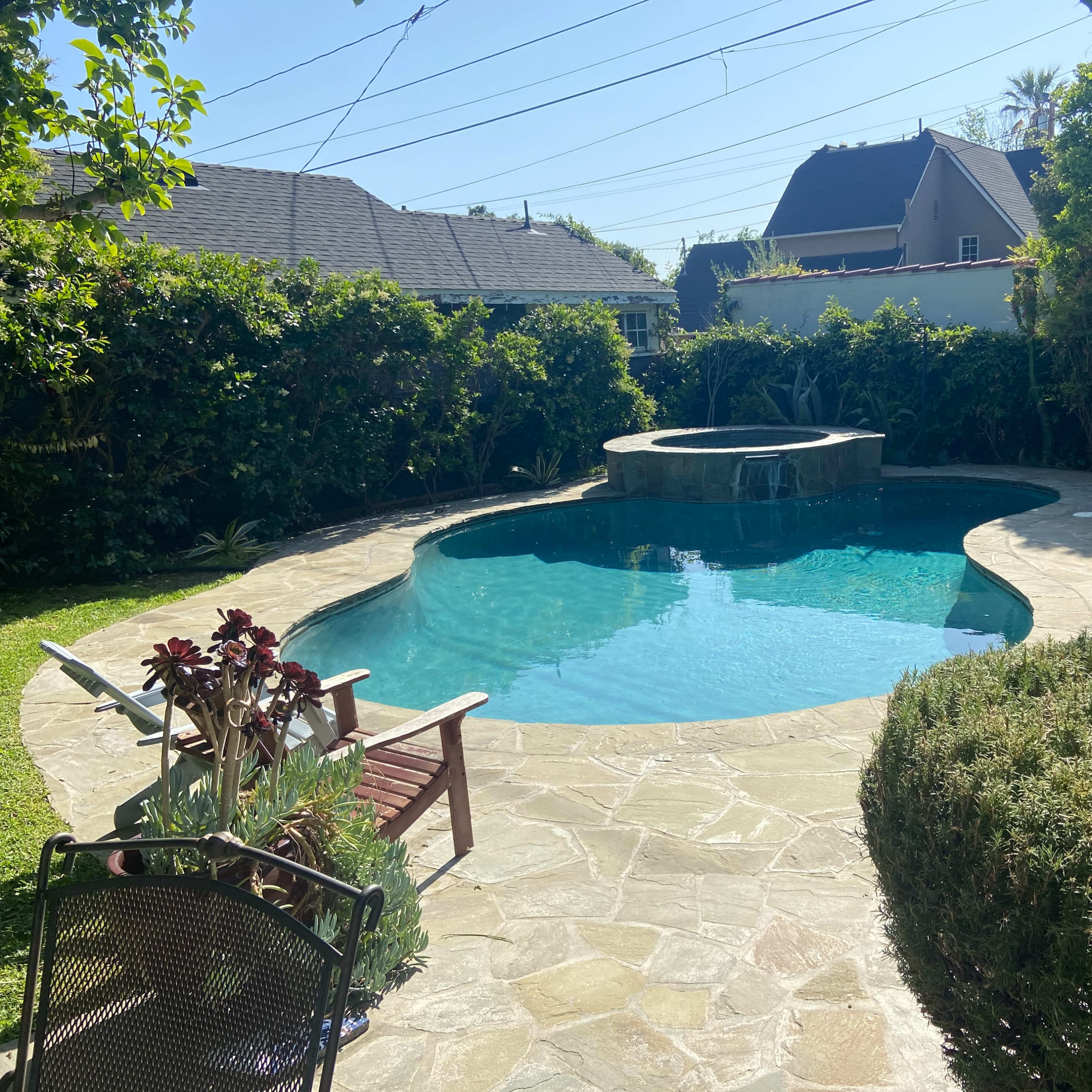 A backyard with a swimming pool featuring a spa, surrounded by lush green landscaping and stonework.