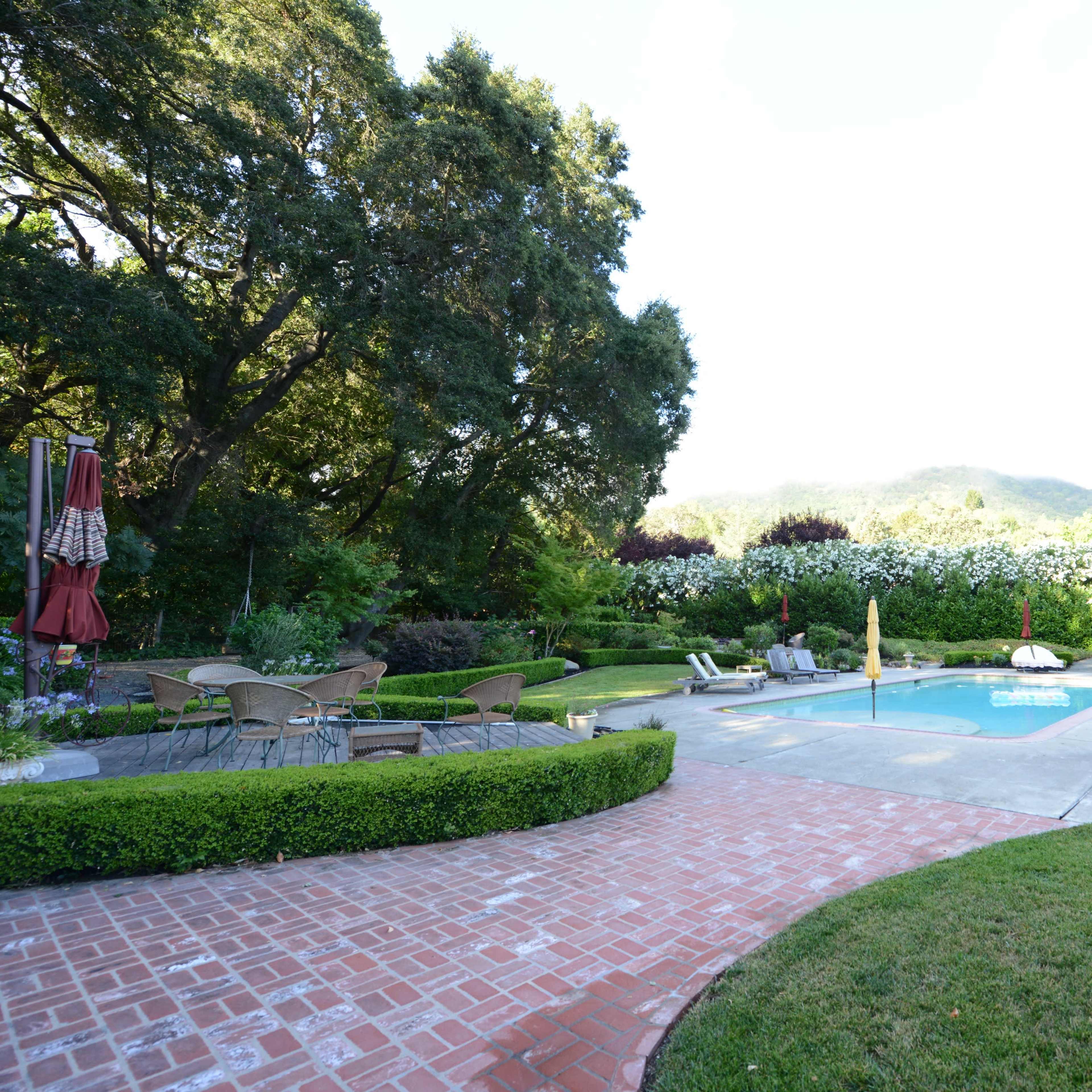 A landscaped backyard featuring a swimming pool surrounded by lounge chairs, a patio area with outdoor furniture, and lush greenery with trees and flowering plants in the background.
