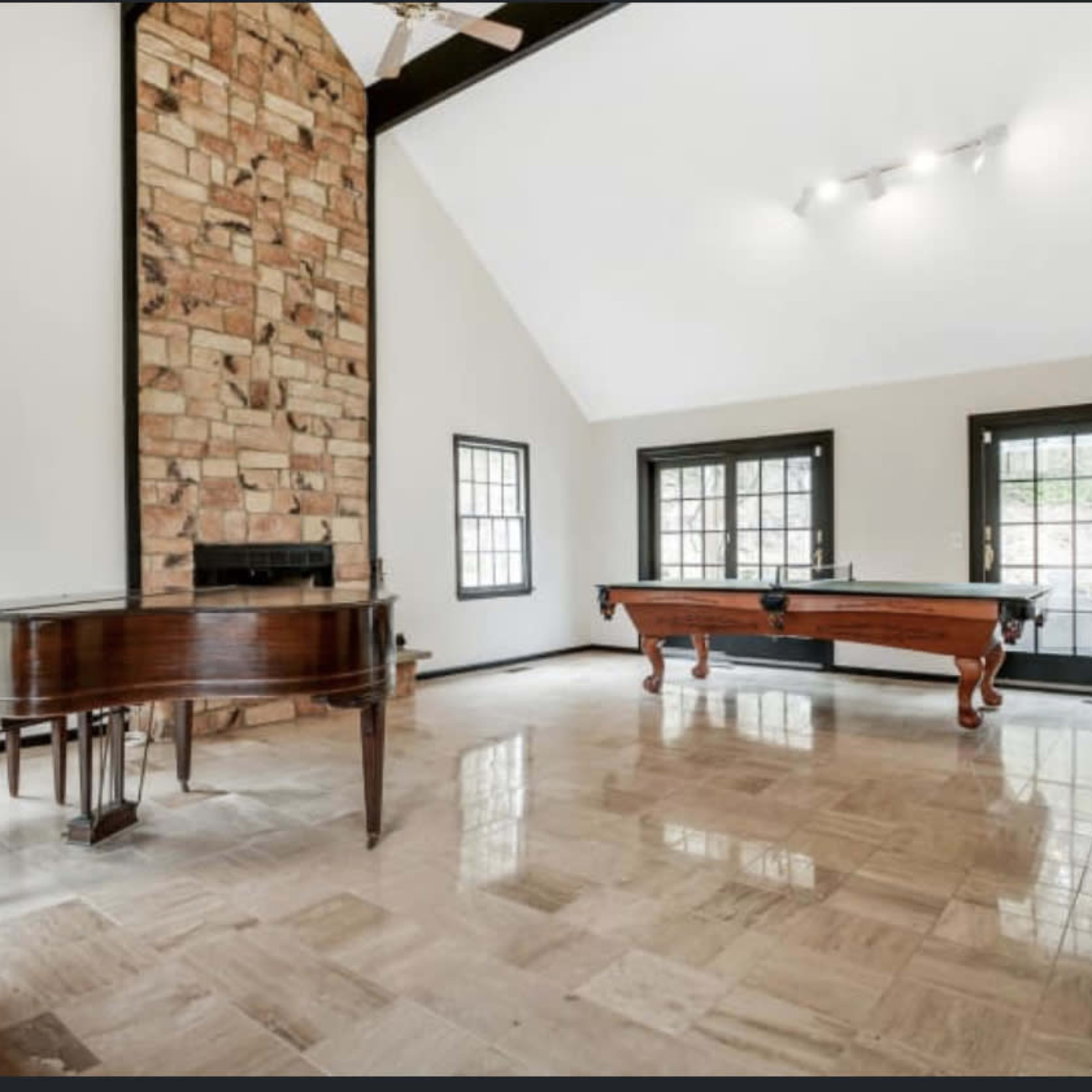 The room features a stone fireplace, a piano, and a billiard table, with large windows allowing natural light to enter.
