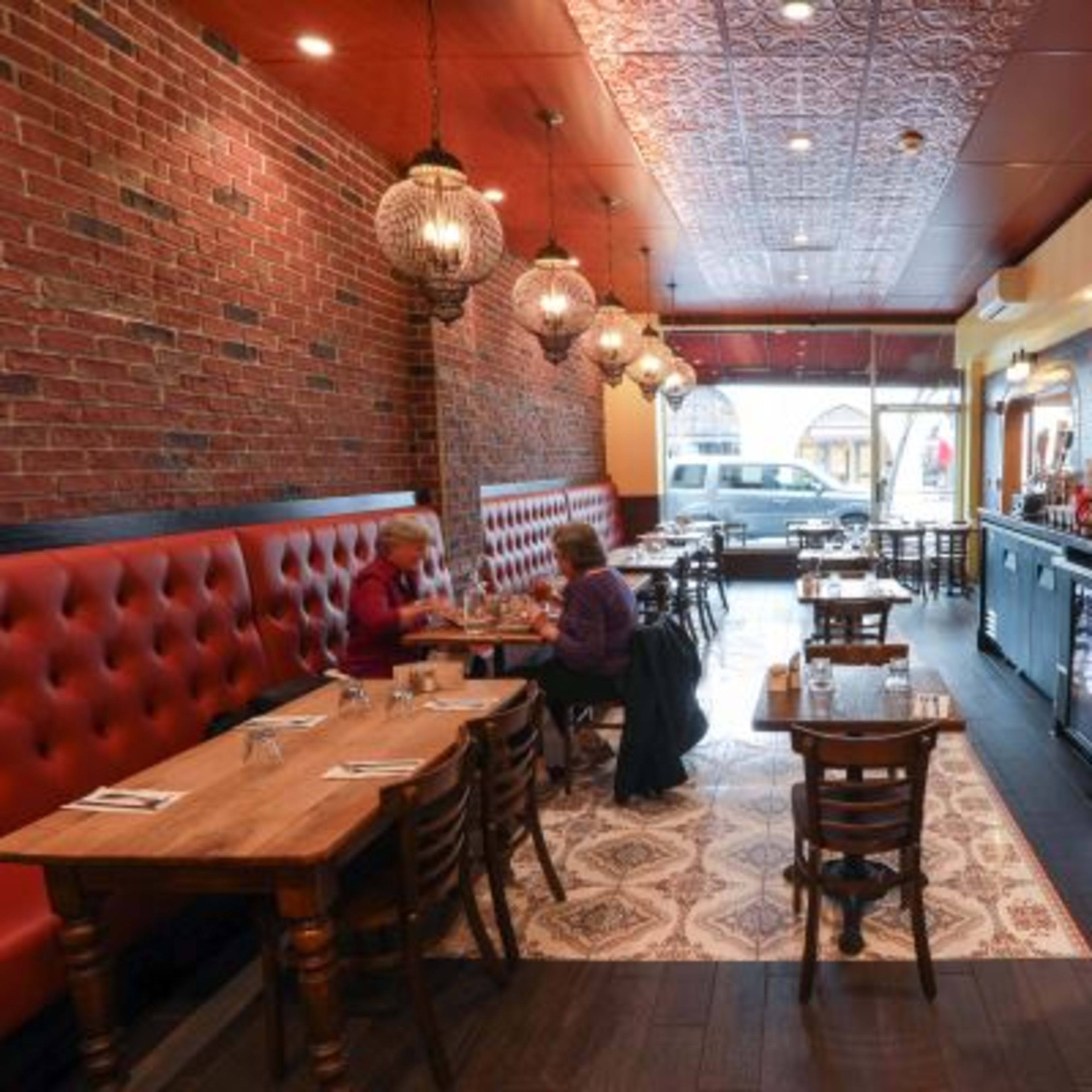 A cozy restaurant interior with wooden tables, red tufted seating, and pendant lights, featuring a brick wall and a serving counter.