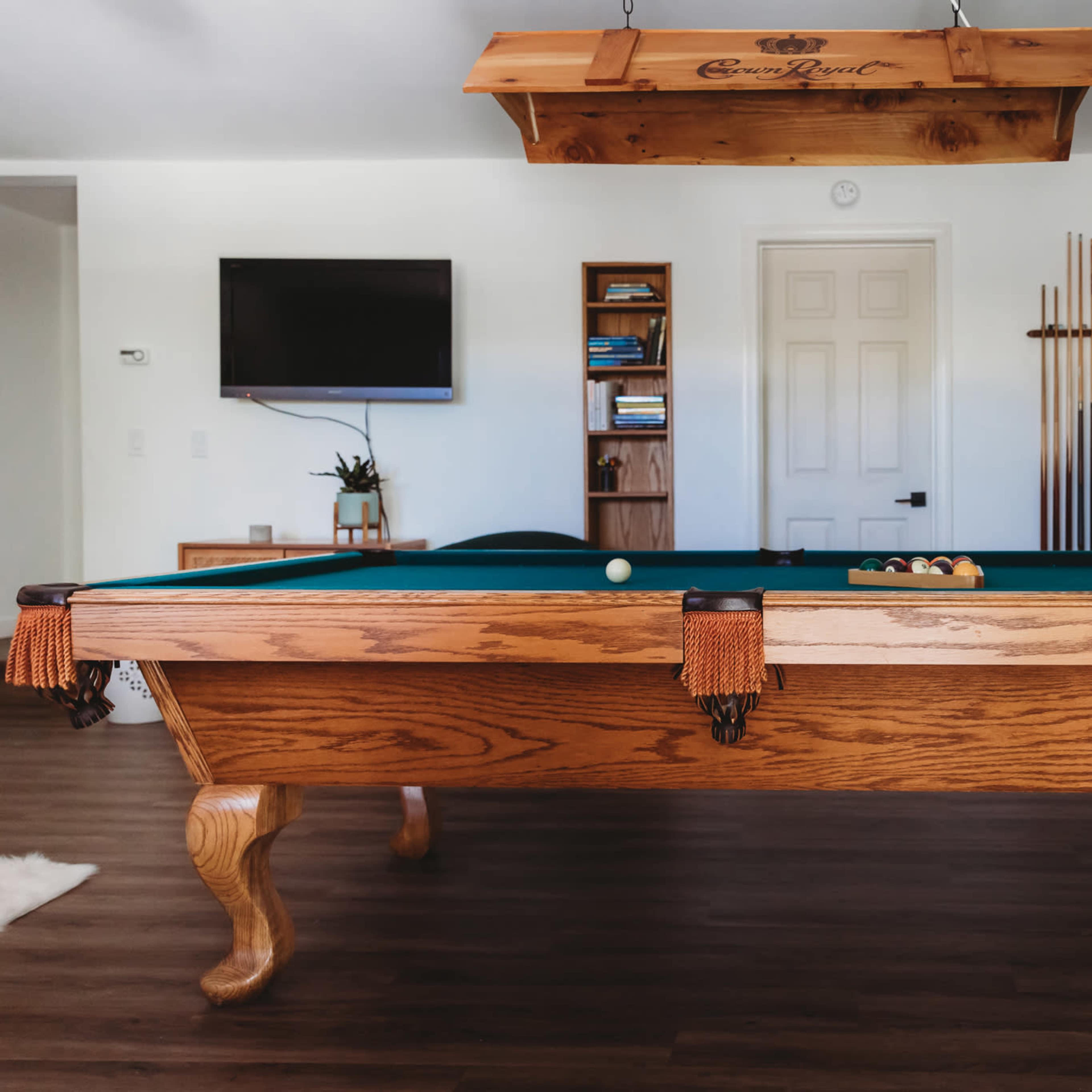 A wooden pool table with green felt, positioned in a well-lit room featuring a television and wall-mounted cues in the background.