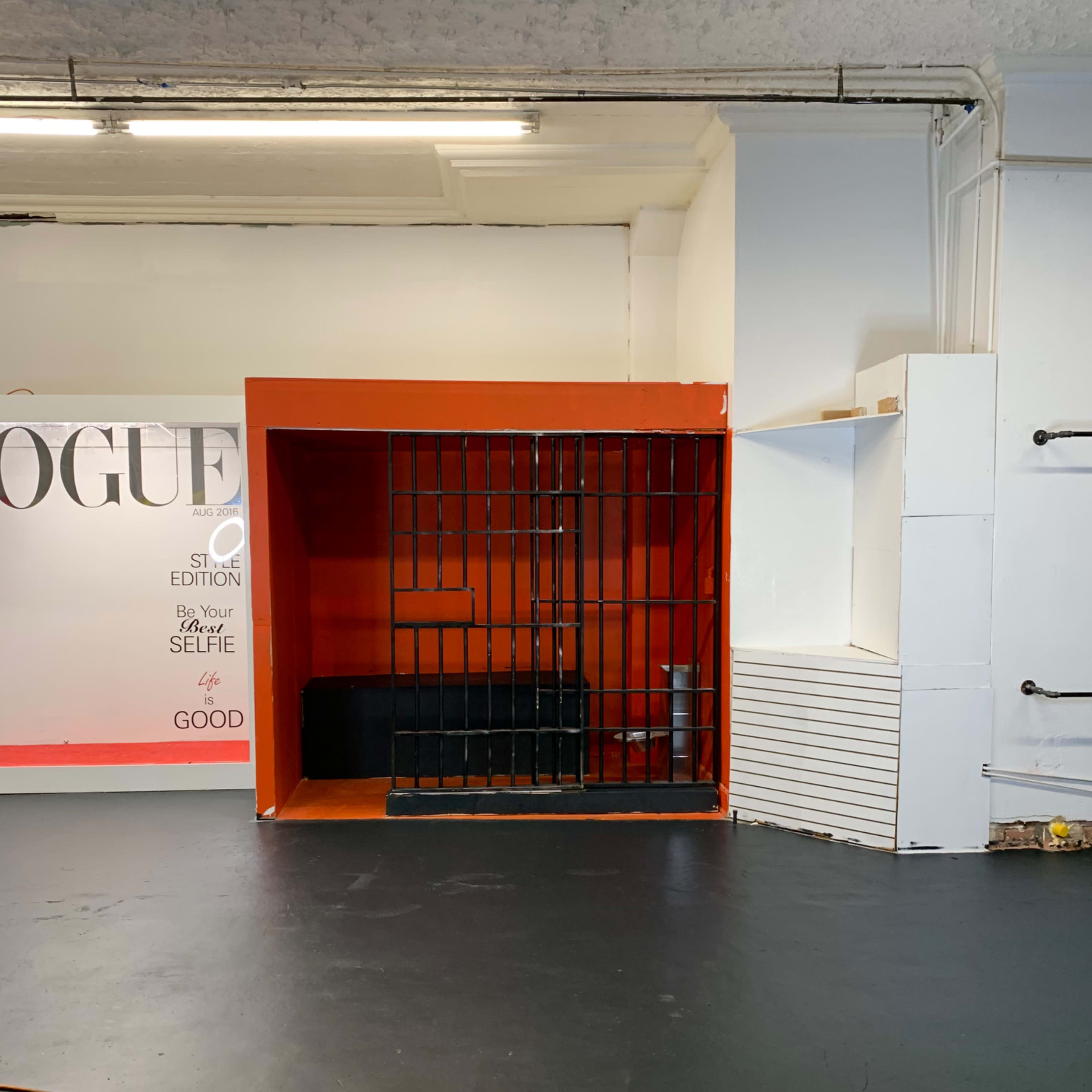 An empty room featuring a red backdrop with a barred enclosure on one side and a white wall with a shelf on the other.
