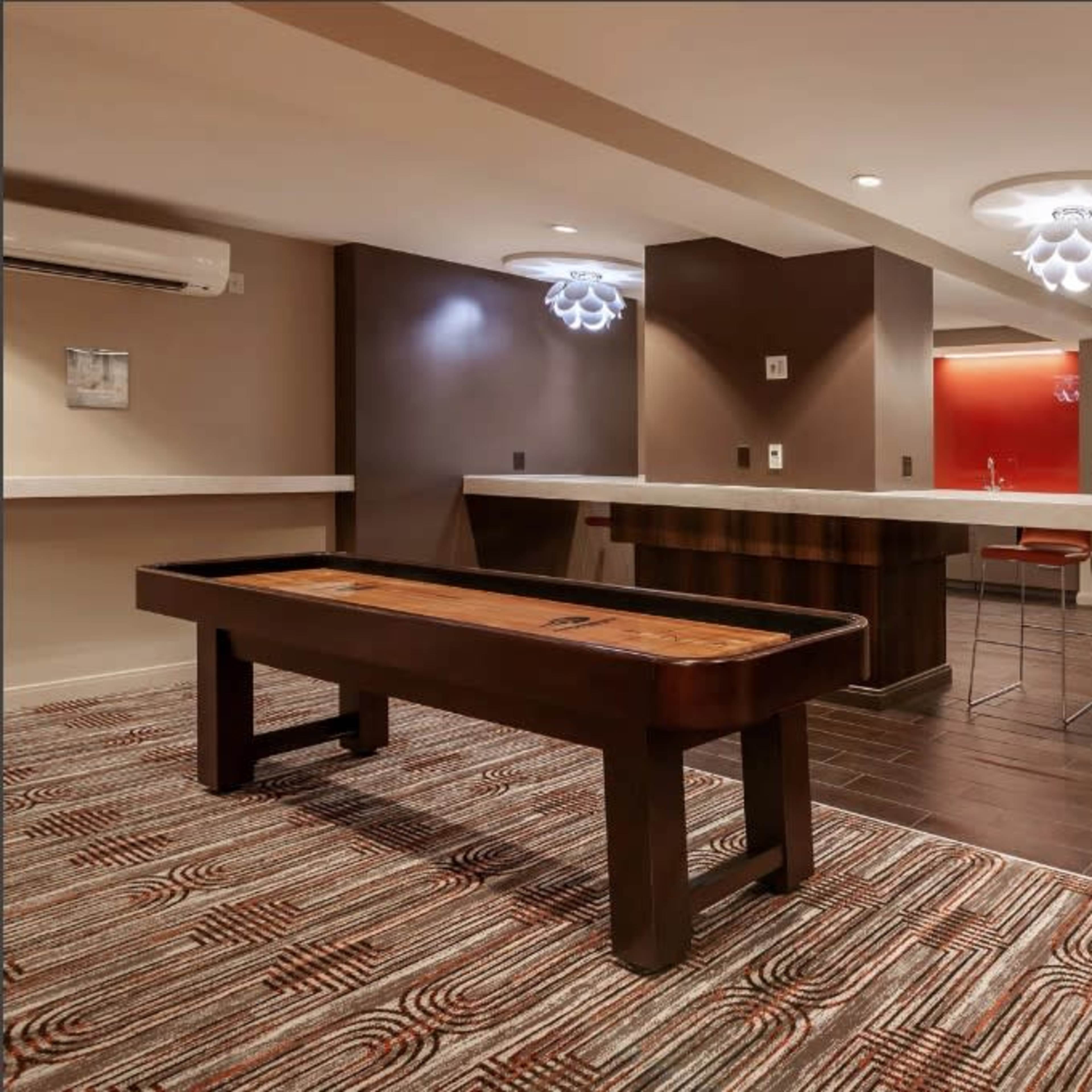 A modern lounge area featuring a shuffleboard table, bar counter, and seating arrangements on polished wood flooring.