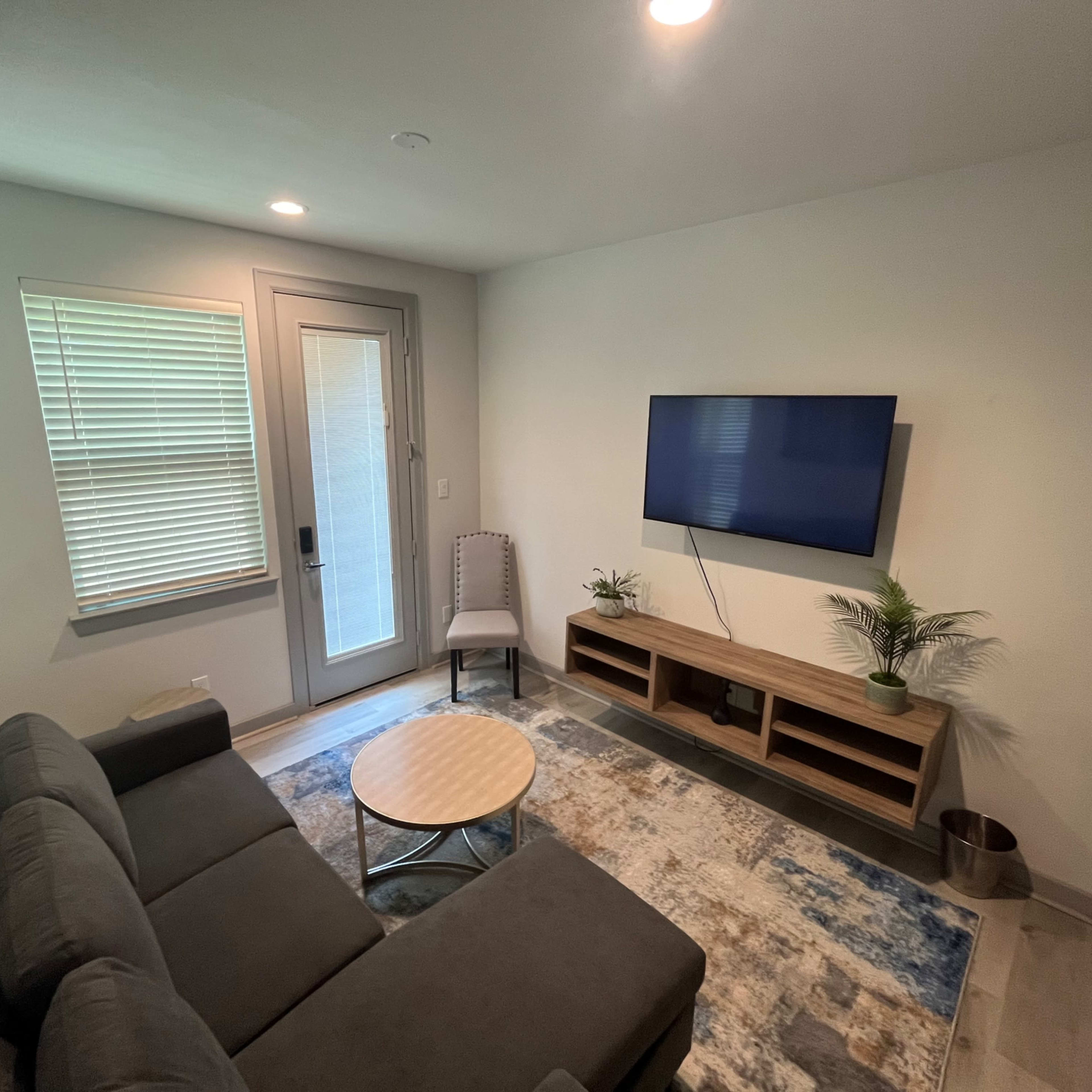The room features a grey sectional sofa, a round coffee table, a television mounted on the wall, and a door leading outside, with a small chair nearby.