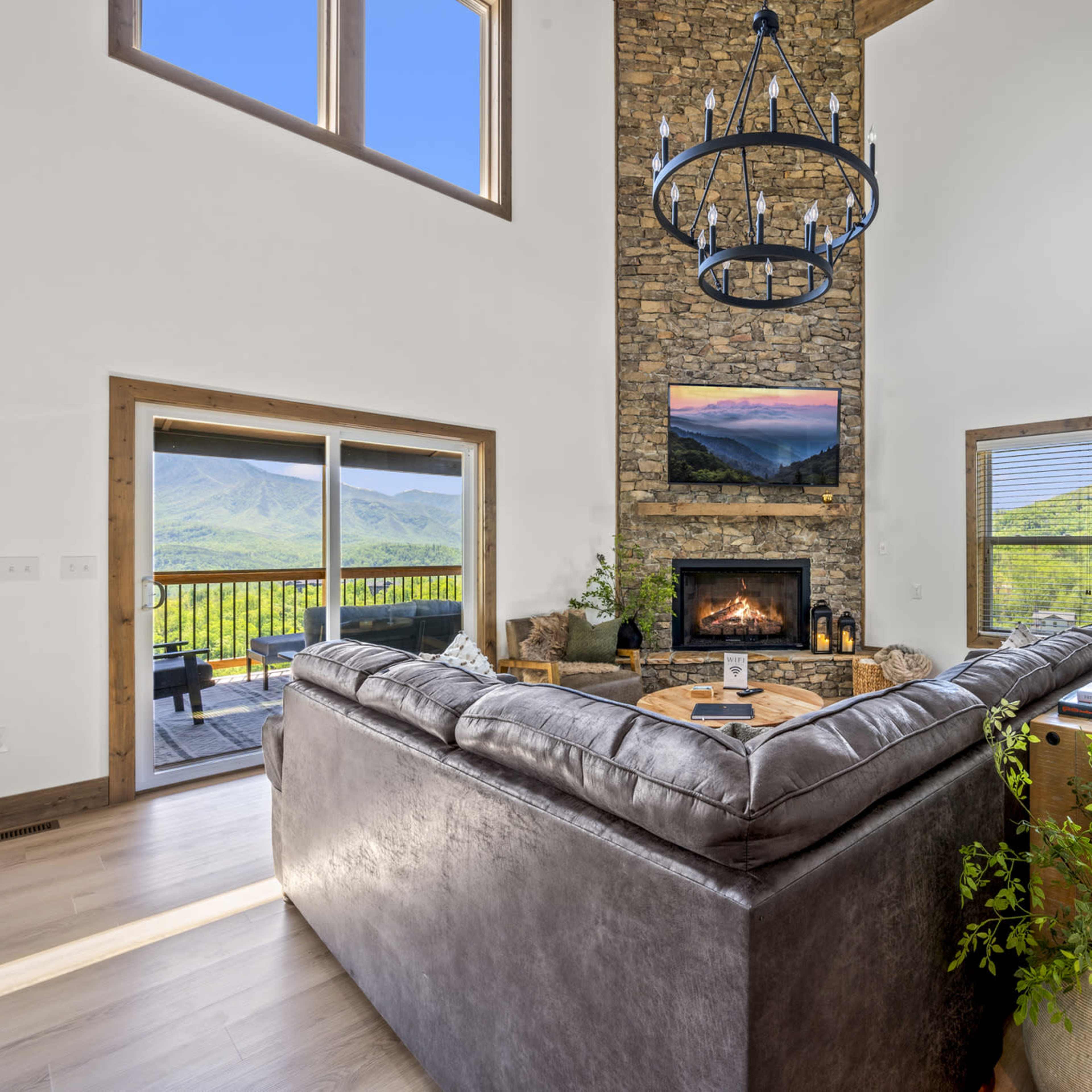 A modern living room with a large sectional sofa facing a stone fireplace, a mounted television above it, and large windows offering a view of the mountains.