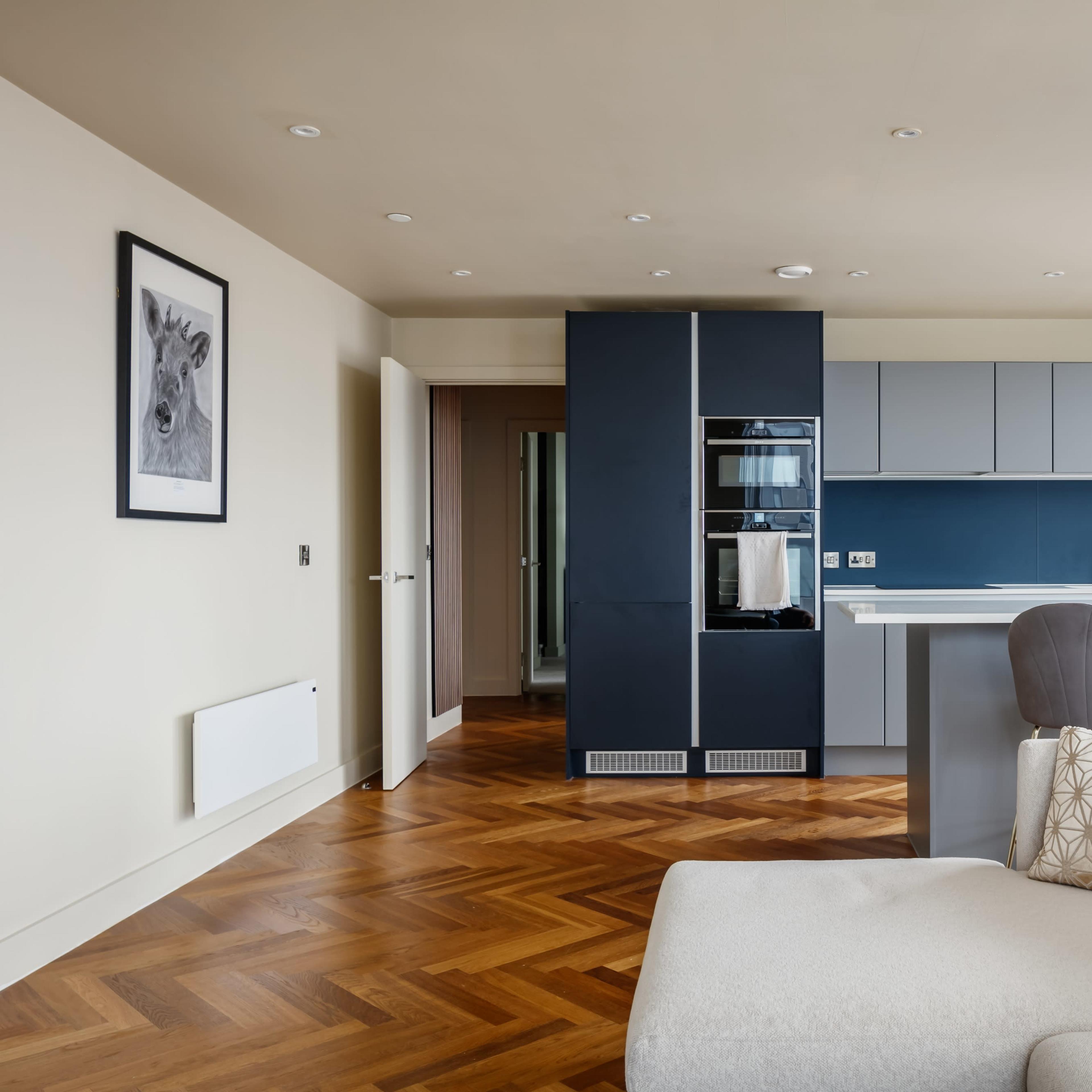 A modern kitchen and living area features a sleek design with a blue kitchen cabinet unit, hardwood flooring, and large windows providing a view of the city.