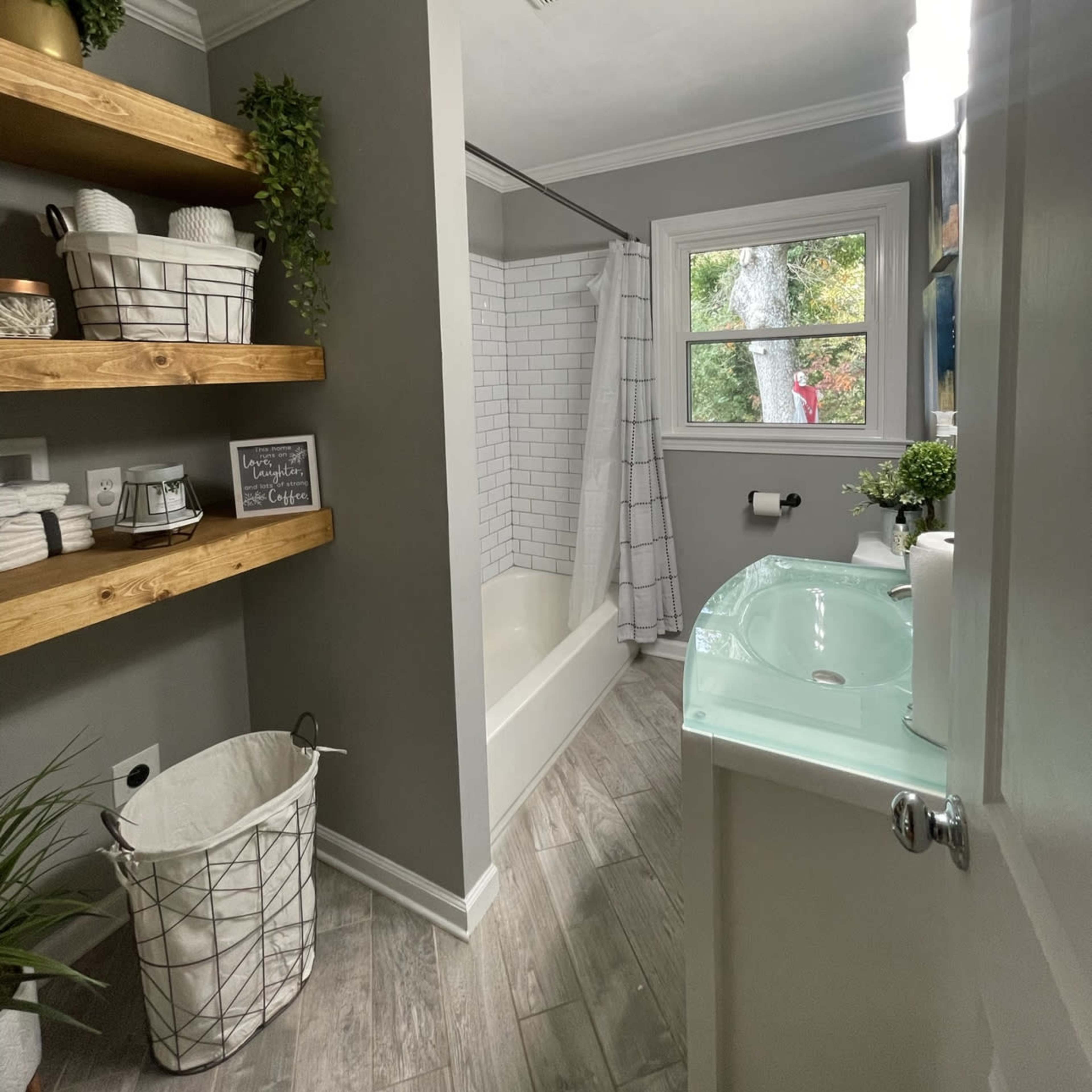 A modern bathroom with a shower-tub combination, wooden shelves holding towels, and a lighted mirror above a sink.
