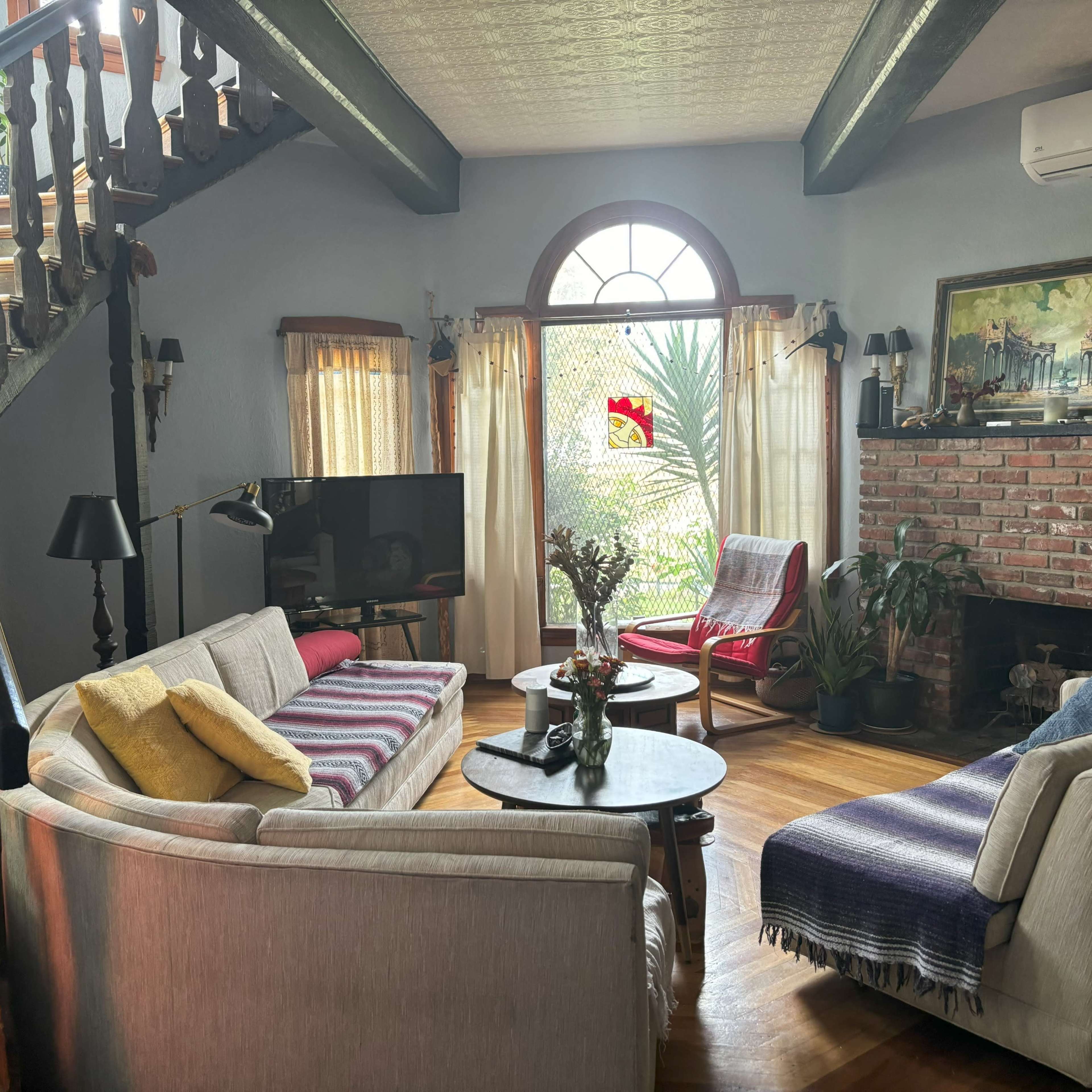 A cozy living room with a staircase, a variety of seating options, a television, and a brick fireplace, all illuminated by natural light from a large window.
