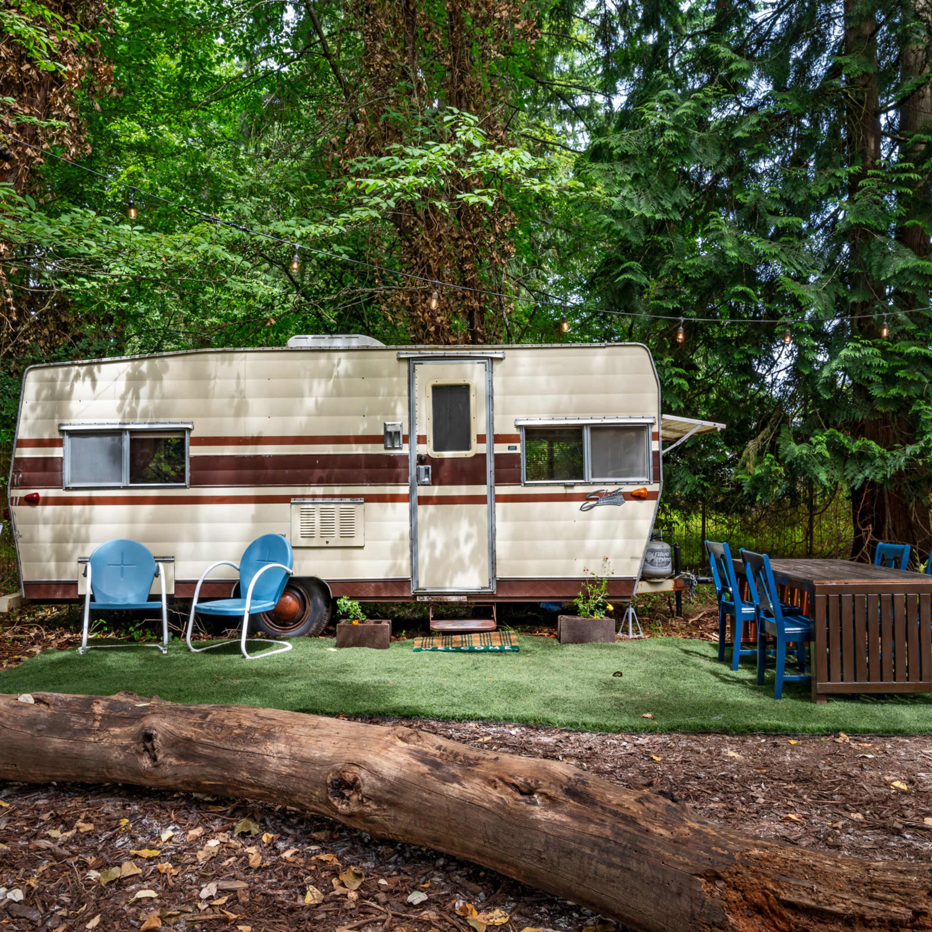 A vintage trailer is parked among tall trees, with a wooden dining table and blue chairs set up on a grassy area nearby.