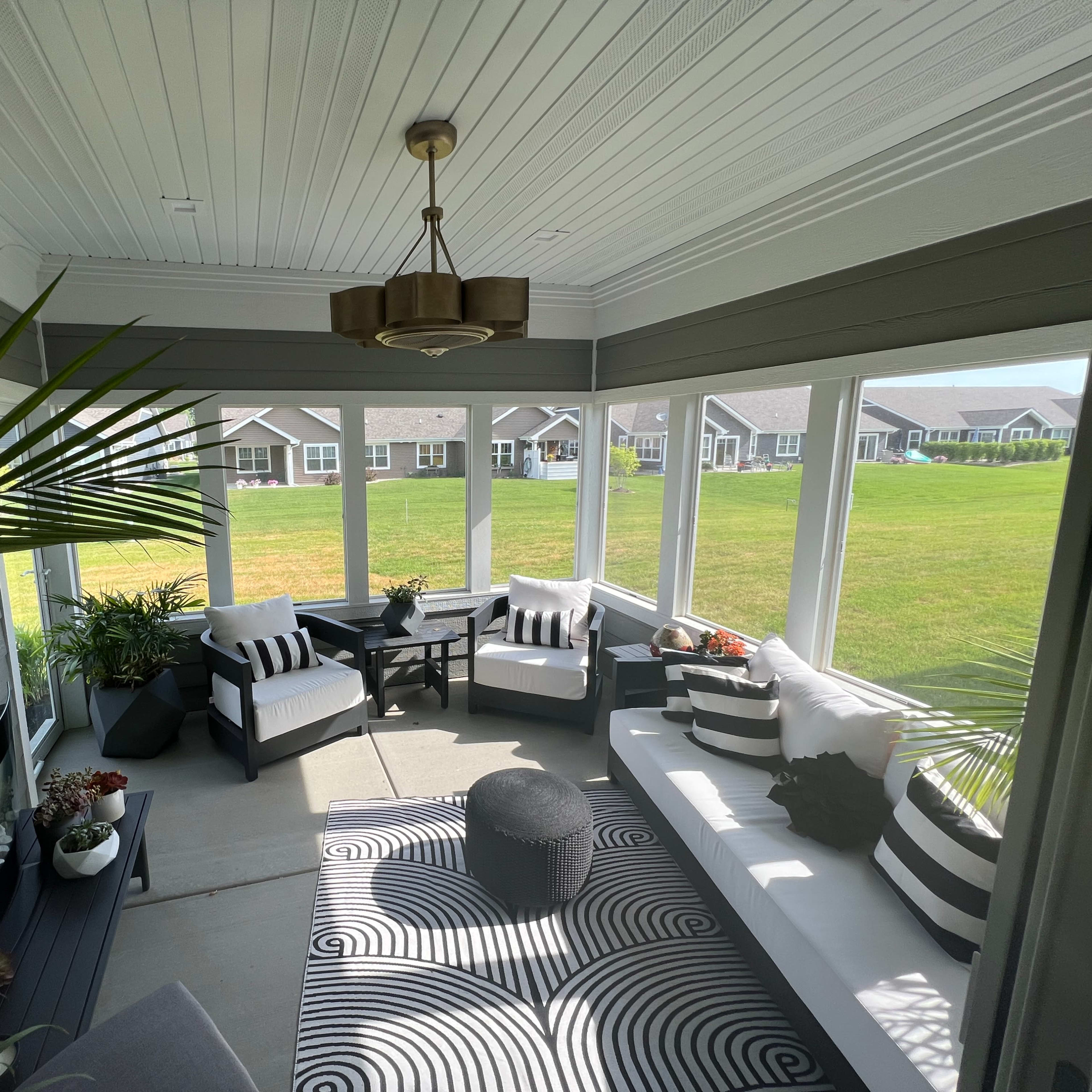 A sunroom with seating furniture, a round table, and indoor plants, featuring large windows overlooking a grassy area.