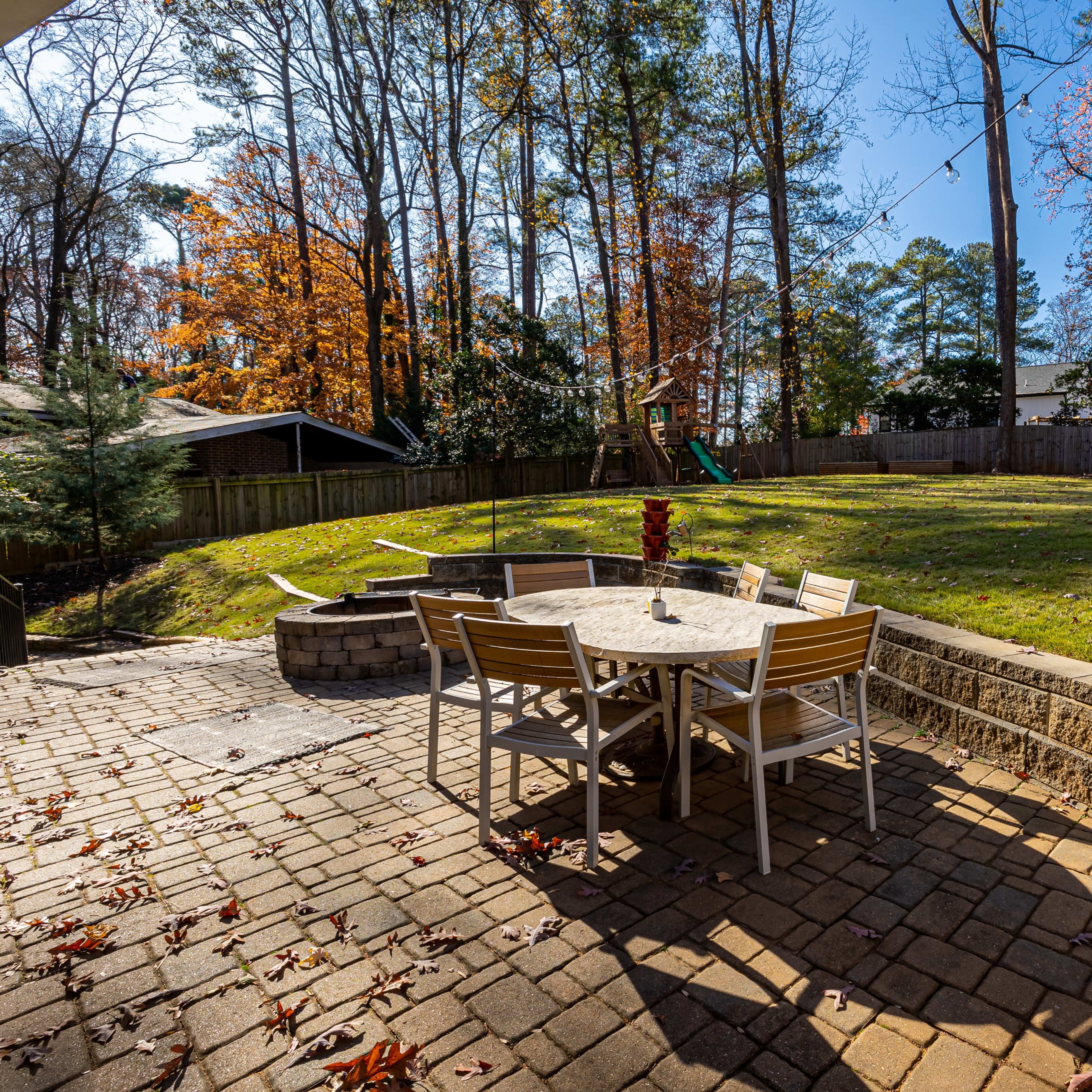 A backyard patio with a stone table and chairs, surrounded by grass and trees with autumn foliage.