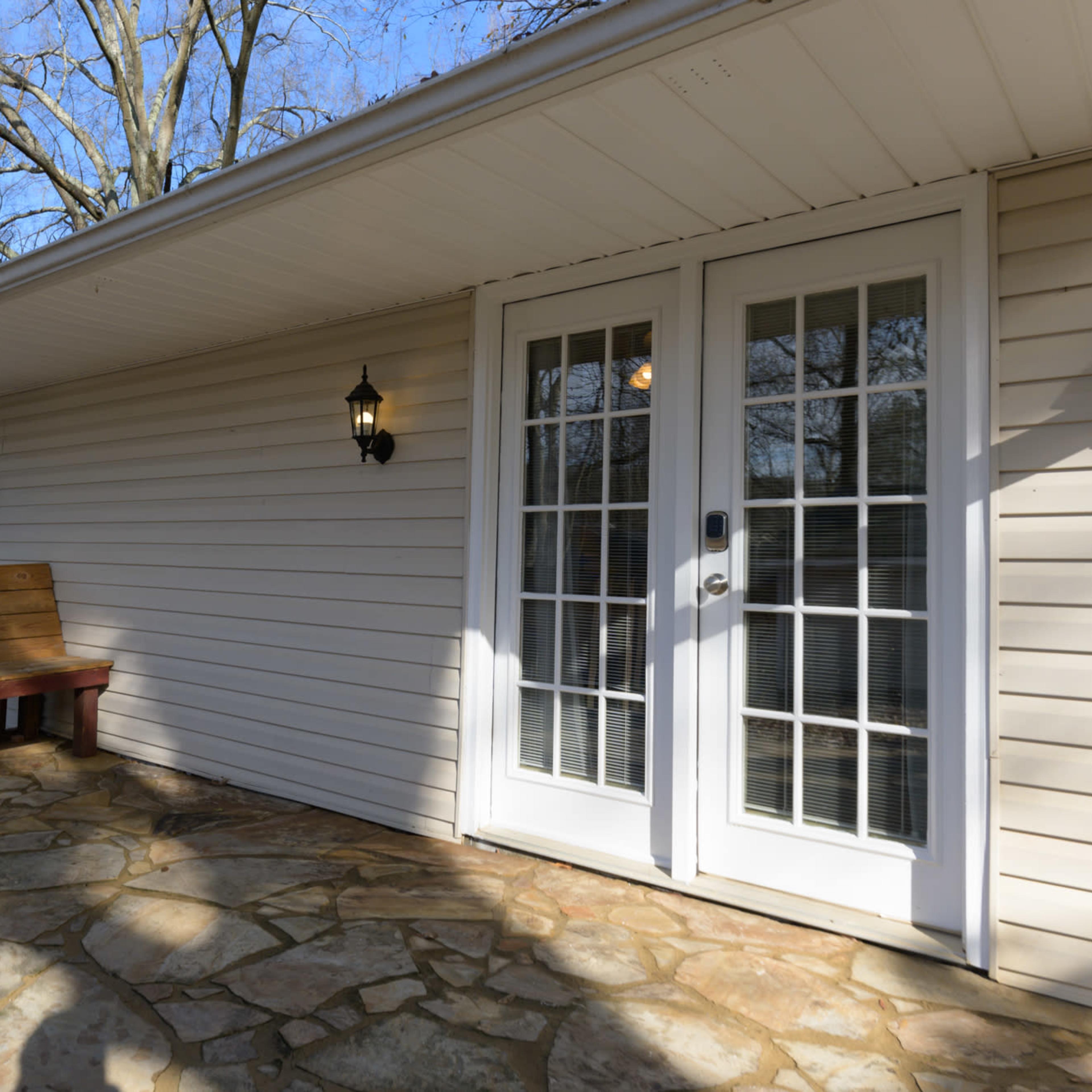 A porch with a stone floor, a wooden bench, and French doors leading into a house.