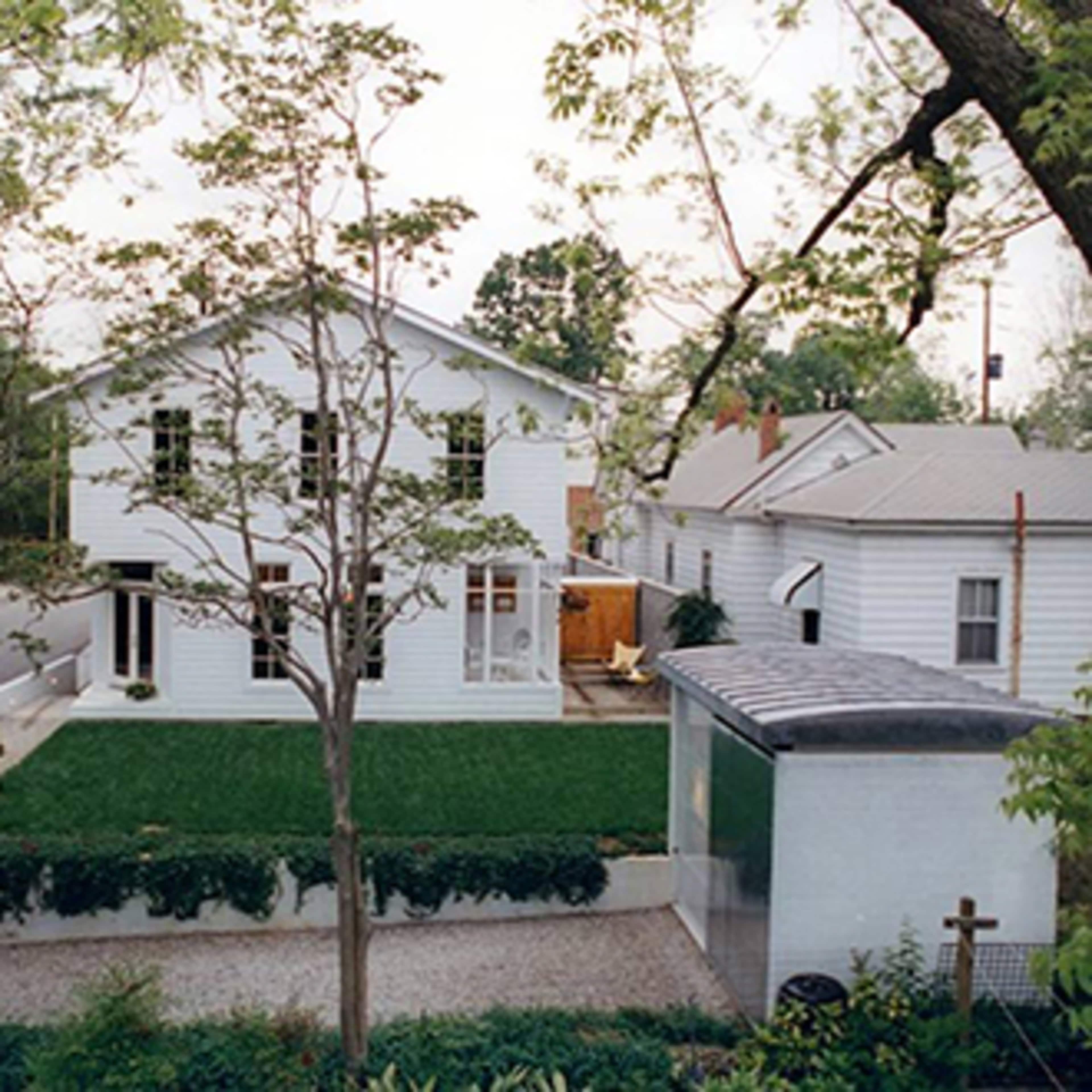 A two-story white house with a green lawn and surrounding trees is pictured, along with adjacent buildings in a residential area.