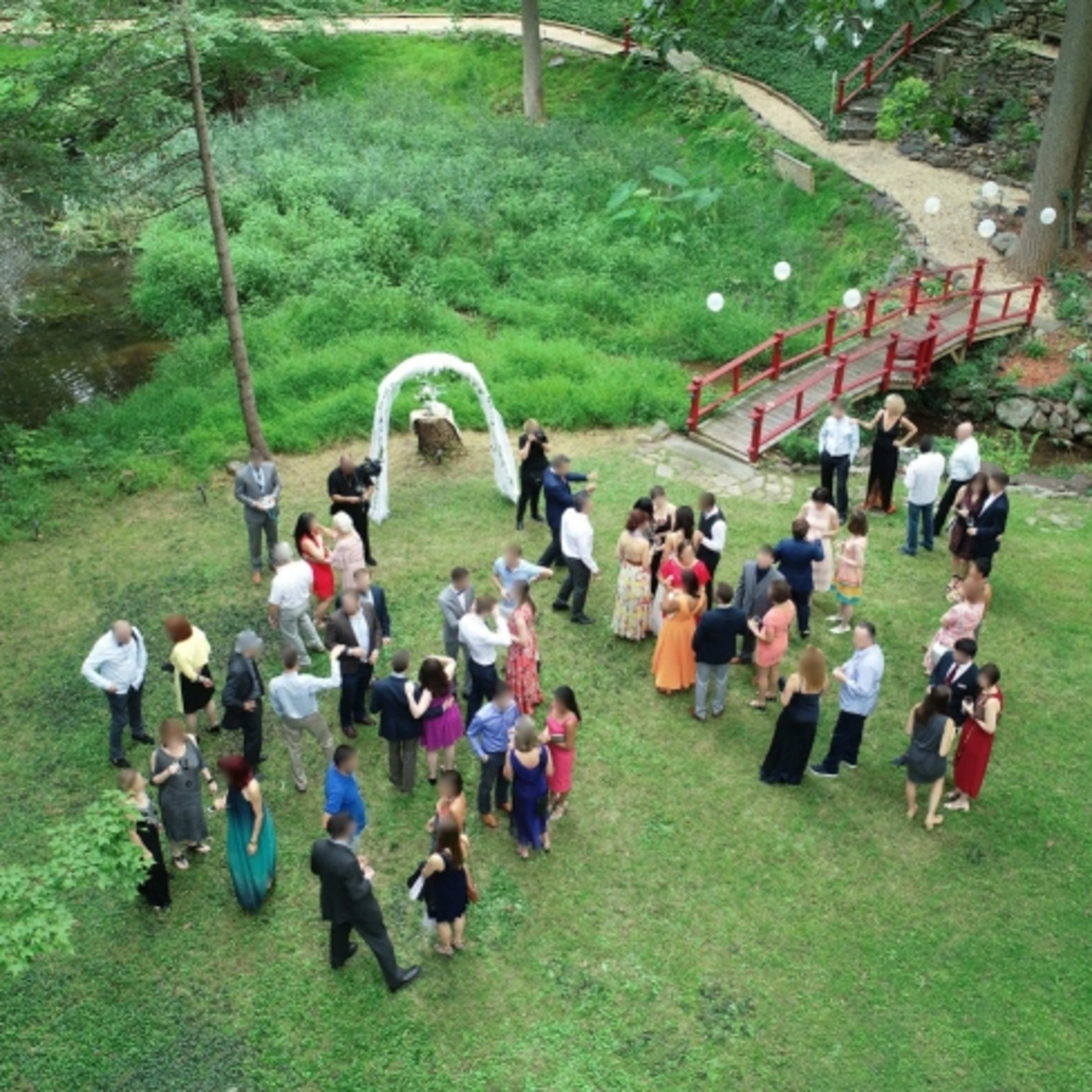 A large outdoor gathering is taking place on a grassy area near a pond, with guests mingling around an arch and a red bridge in the background.